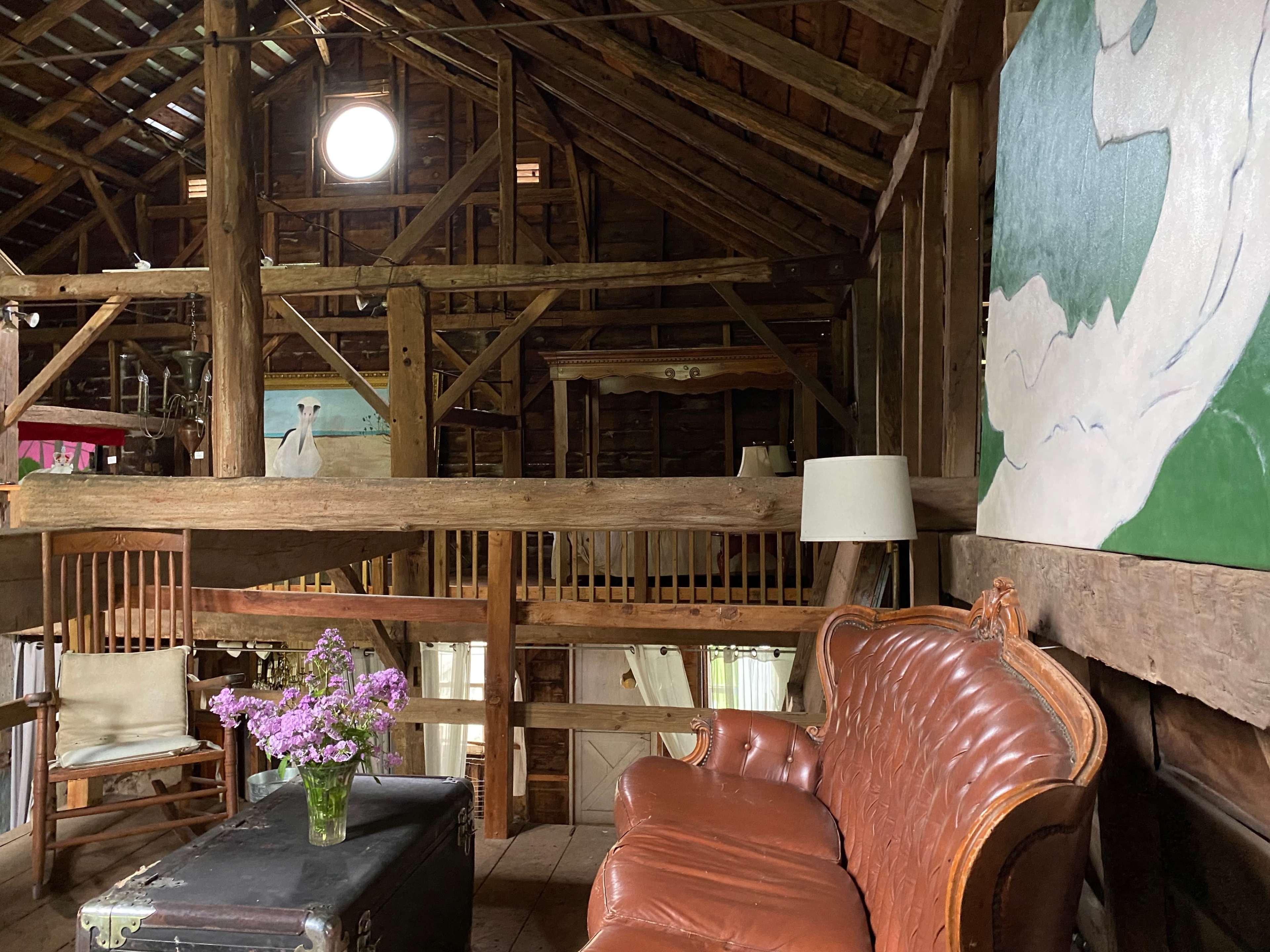 The interior of a rustic barn features wooden beams, a vintage leather sofa, and a vase of flowers on an ornate table, with large paintings on the walls.