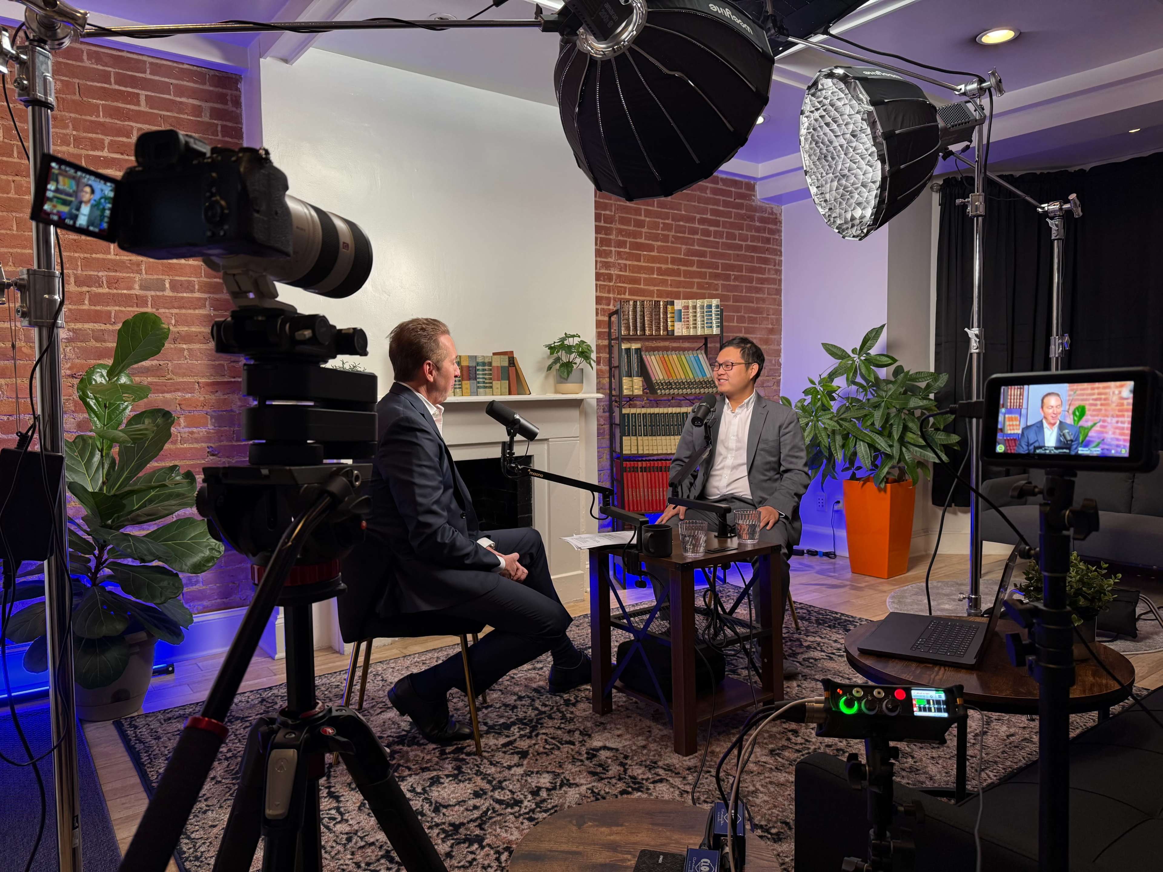 A well-lit interview setup features two men sitting at a table surrounded by cameras, lighting equipment, and indoor plants in a cozy room with a brick wall.