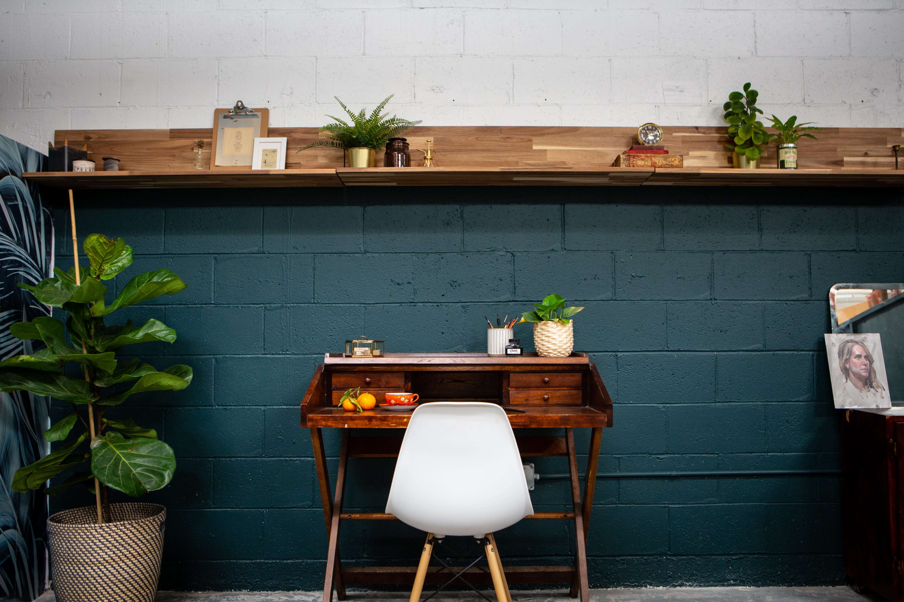 A wooden desk with a white chair is positioned against a dark green wall, accented by a wooden shelf above featuring plants and decorative items.