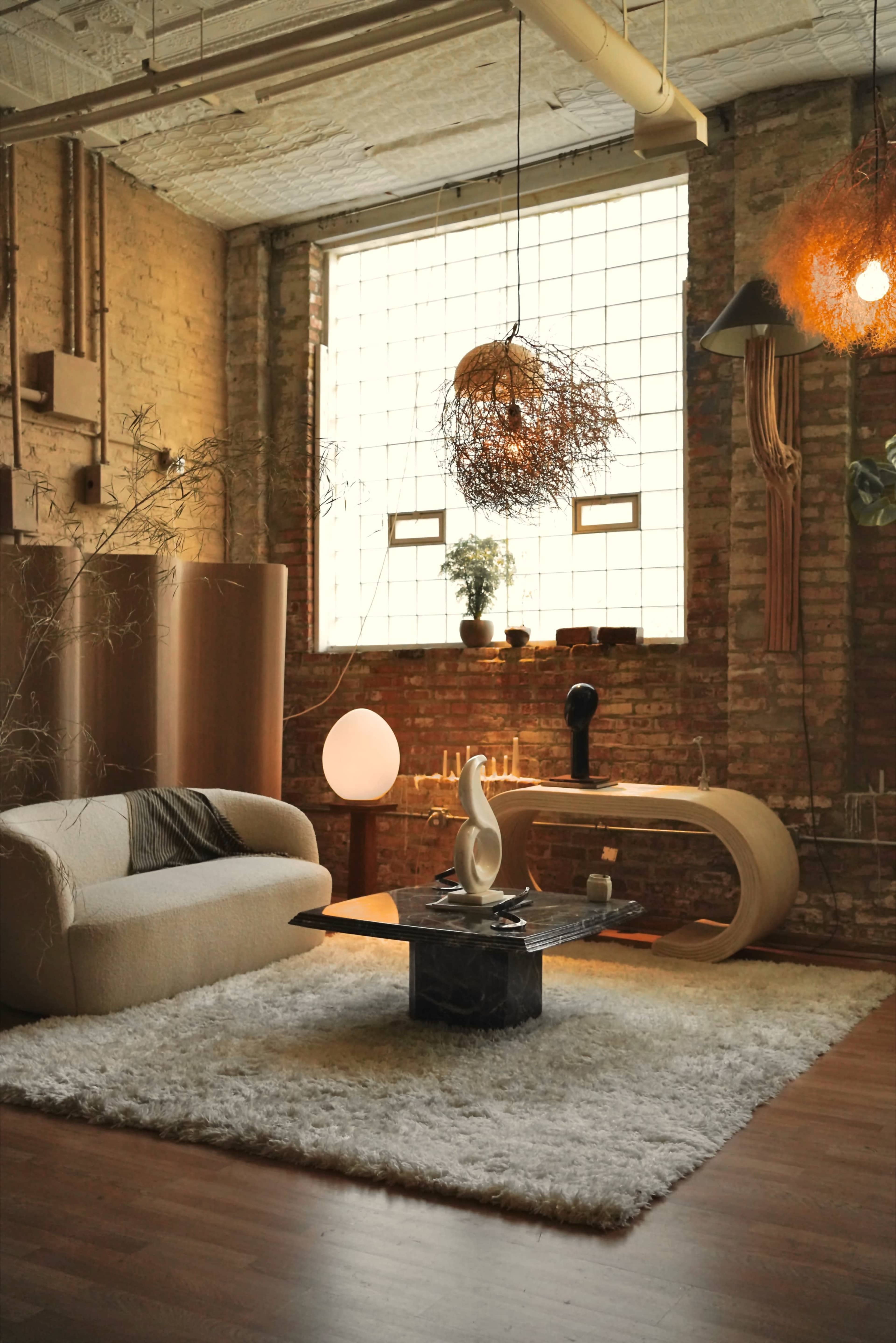 A cozy living room with a large window, a light-colored sofa, a curved wooden coffee table, and a textured rug on a wooden floor.