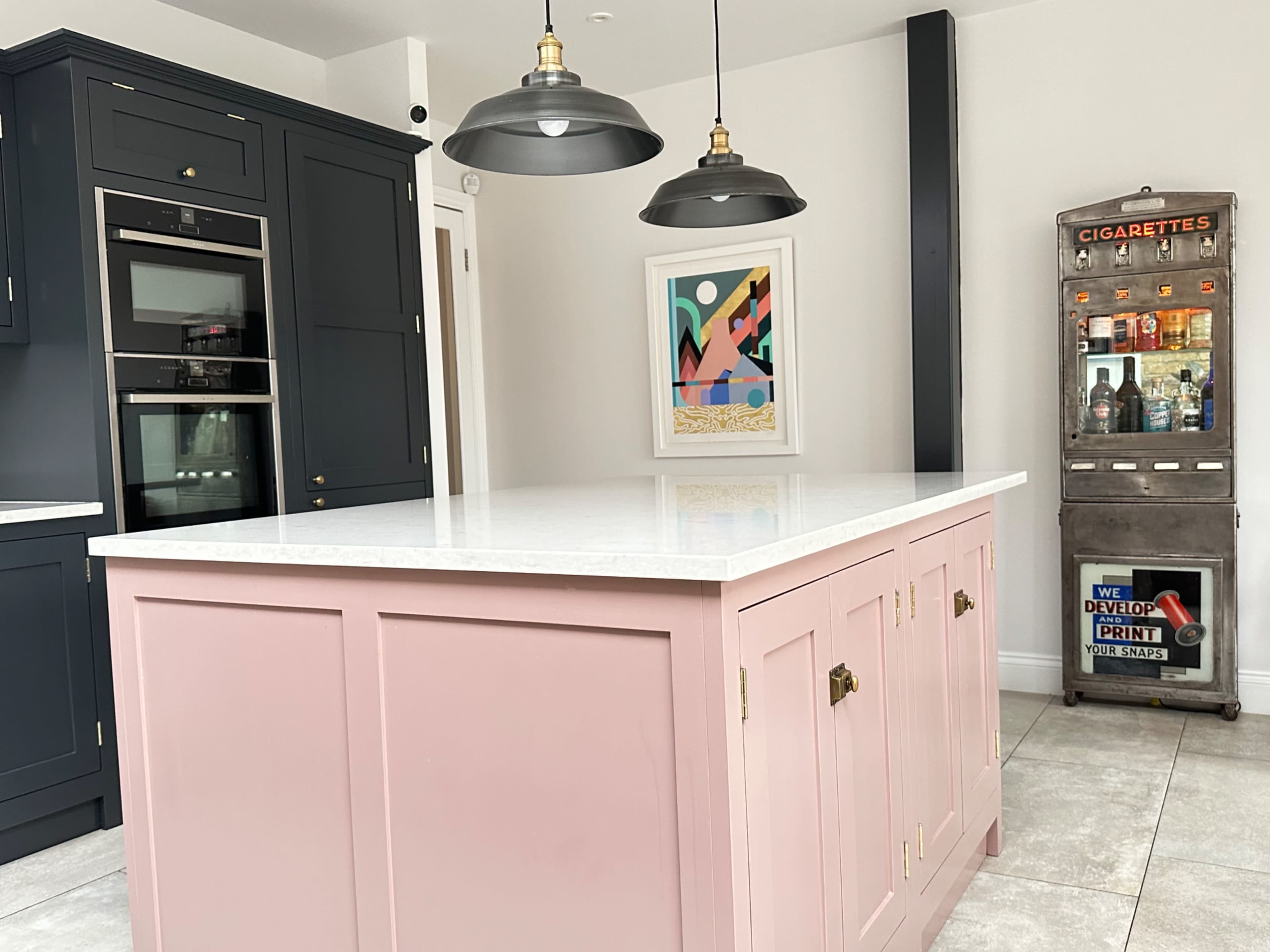 The image shows a modern kitchen with a pink island, dark cabinetry, and pendant lights, alongside an antique cigarette vending machine.