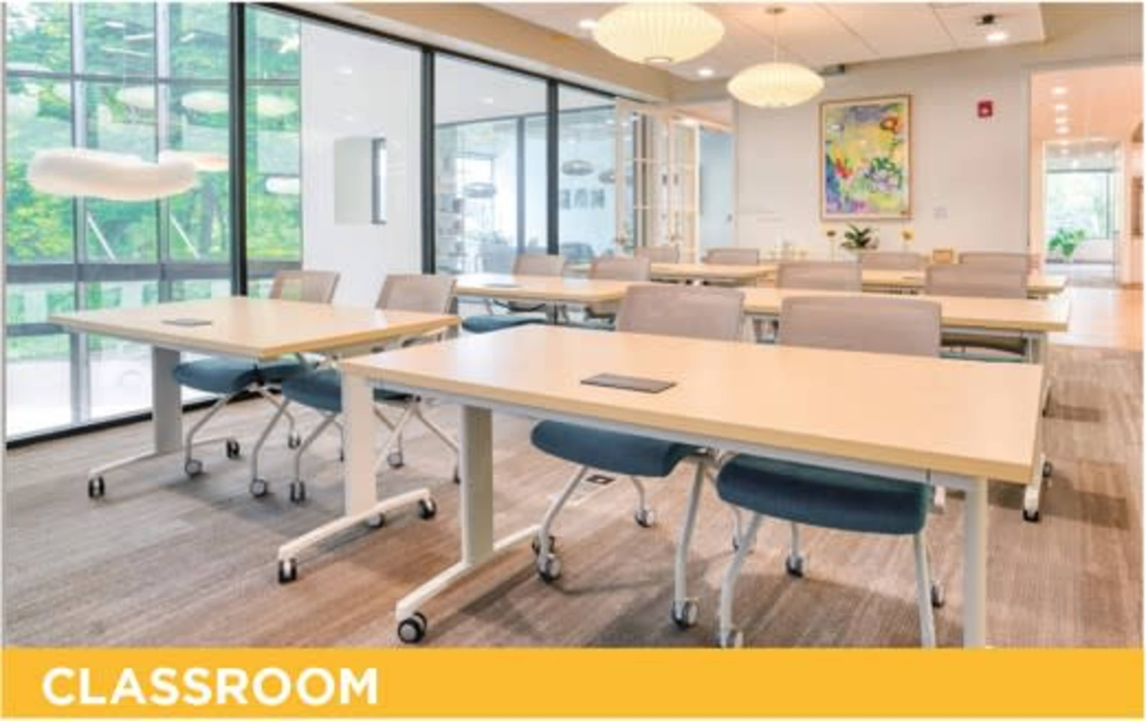 A modern classroom features several tables with rolling chairs arranged neatly, surrounded by large windows that let in natural light.