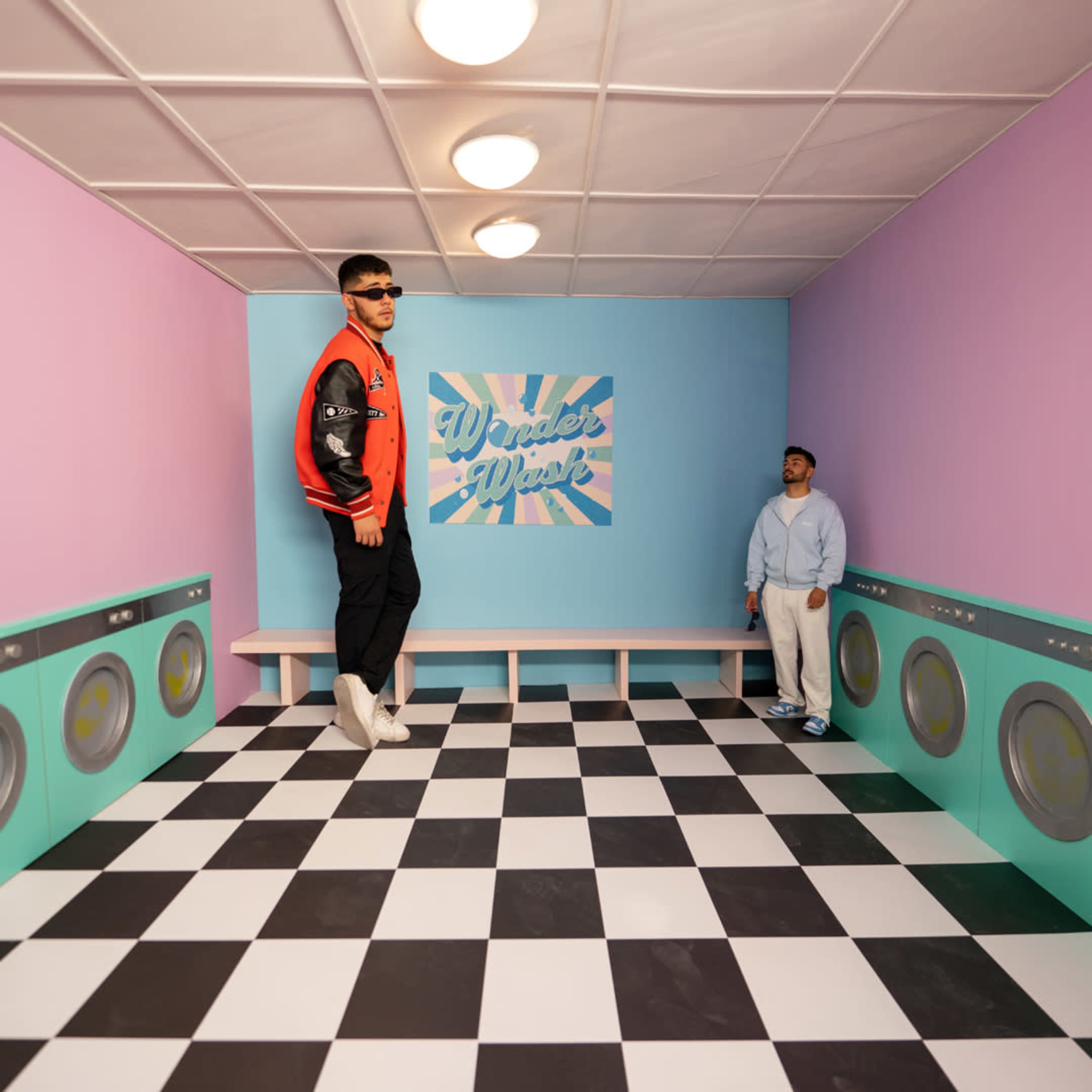 Two individuals are posed in a colorful room designed with a checkered floor and walls in pastel colors, featuring a graphic sign that reads "Wonder Wall."