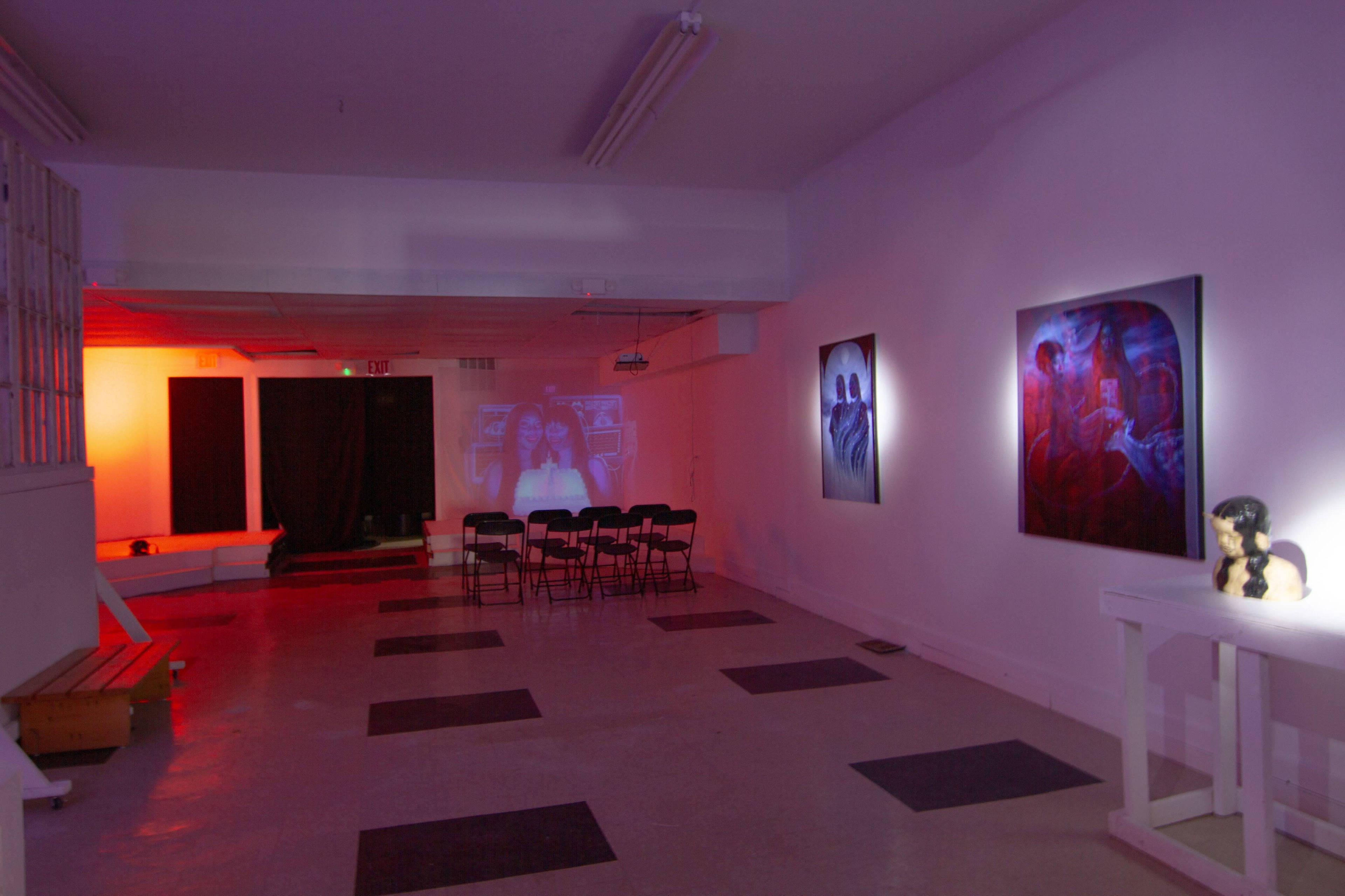 The image shows an art gallery space featuring dim lighting, several artworks on the walls, and a row of empty chairs facing a black curtain.