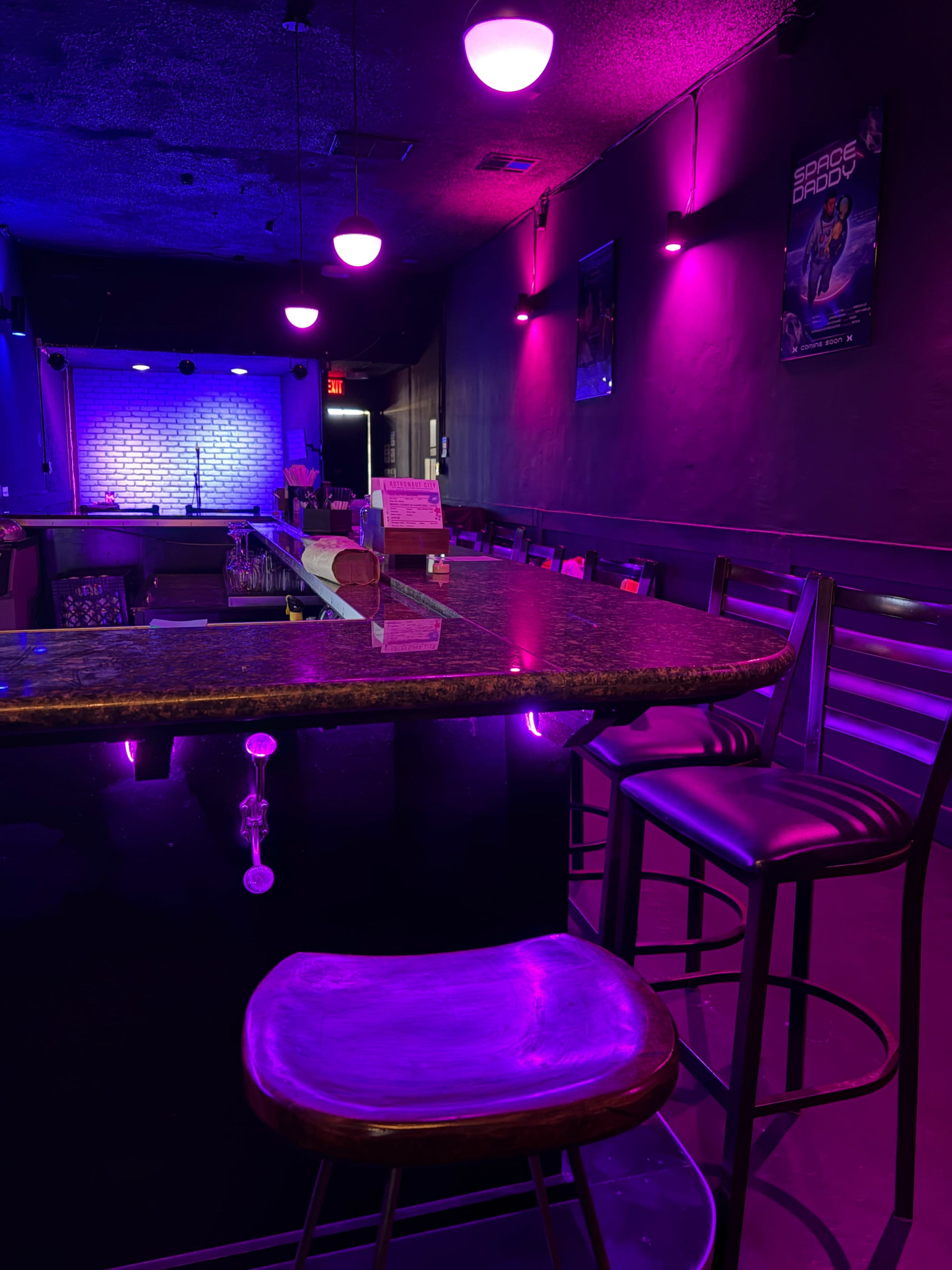 The image shows a dimly lit bar with purple and blue lighting, featuring a polished countertop and bar stools arranged neatly in front of a small stage area.