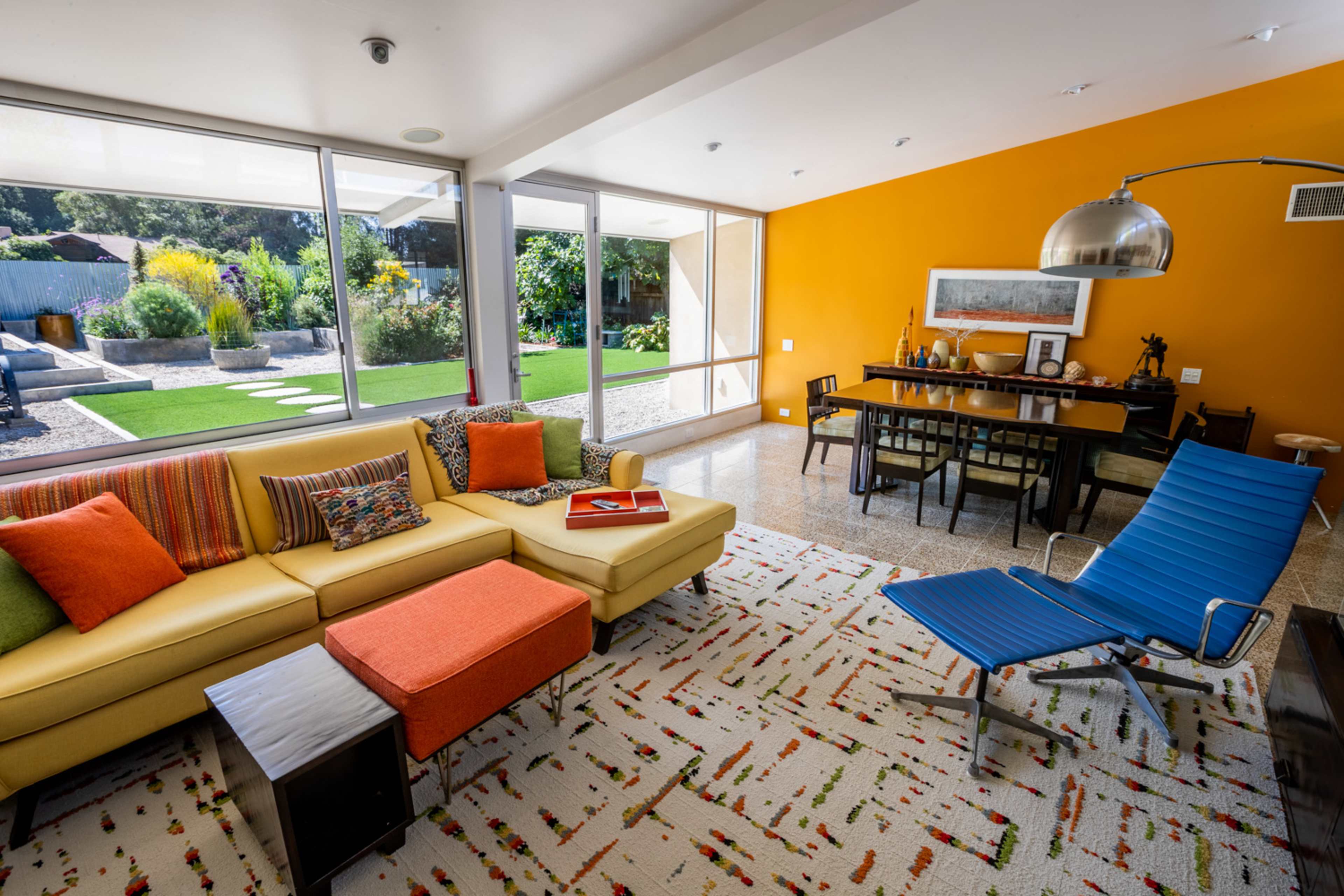 A bright living room with a yellow sofa, an orange ottoman, and a blue lounge chair, overlooking a grassy outdoor area through large windows.