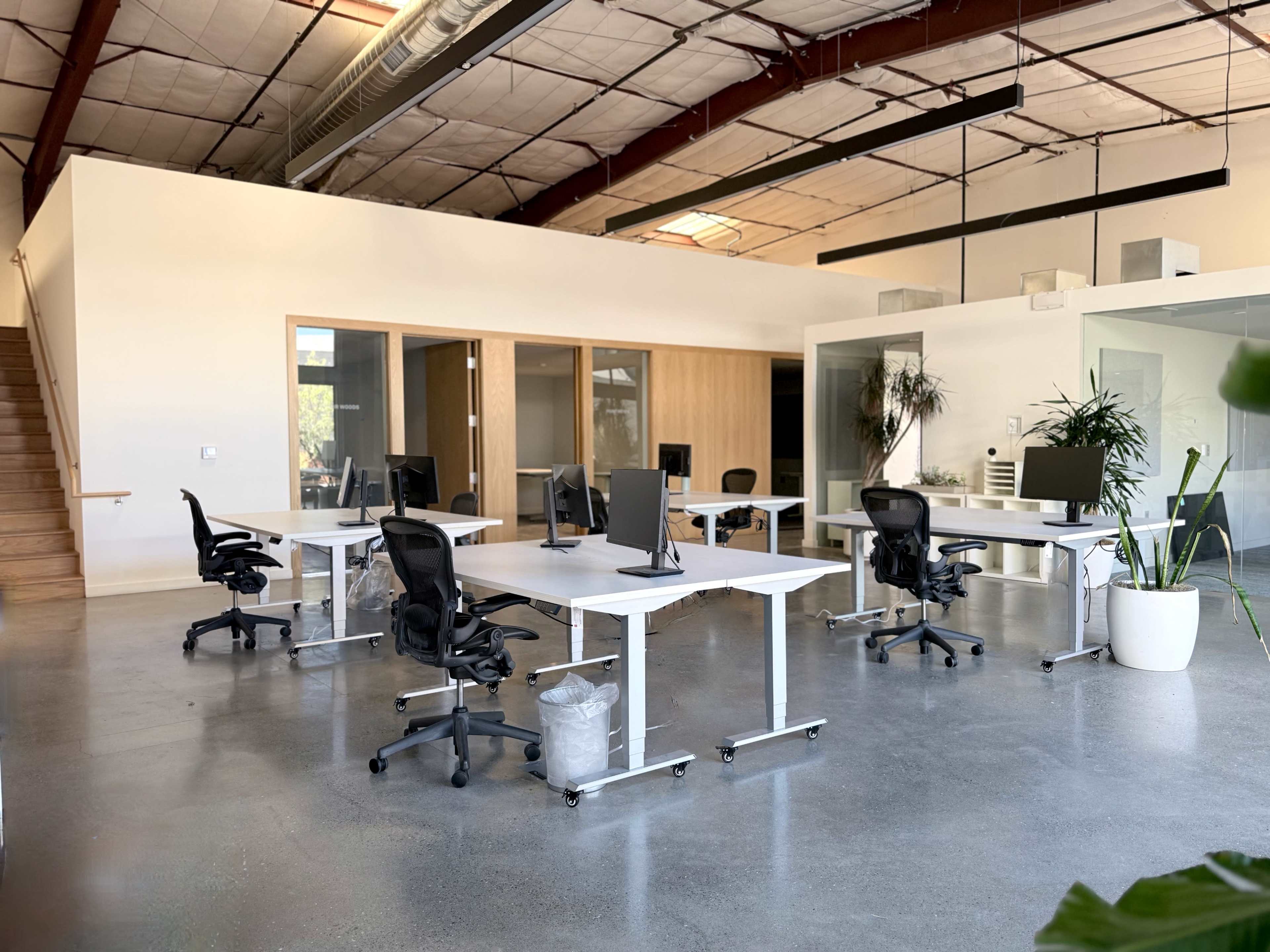 The image shows a modern office space with several desks and chairs arranged in an open layout, featuring large windows and plants for a fresh atmosphere.