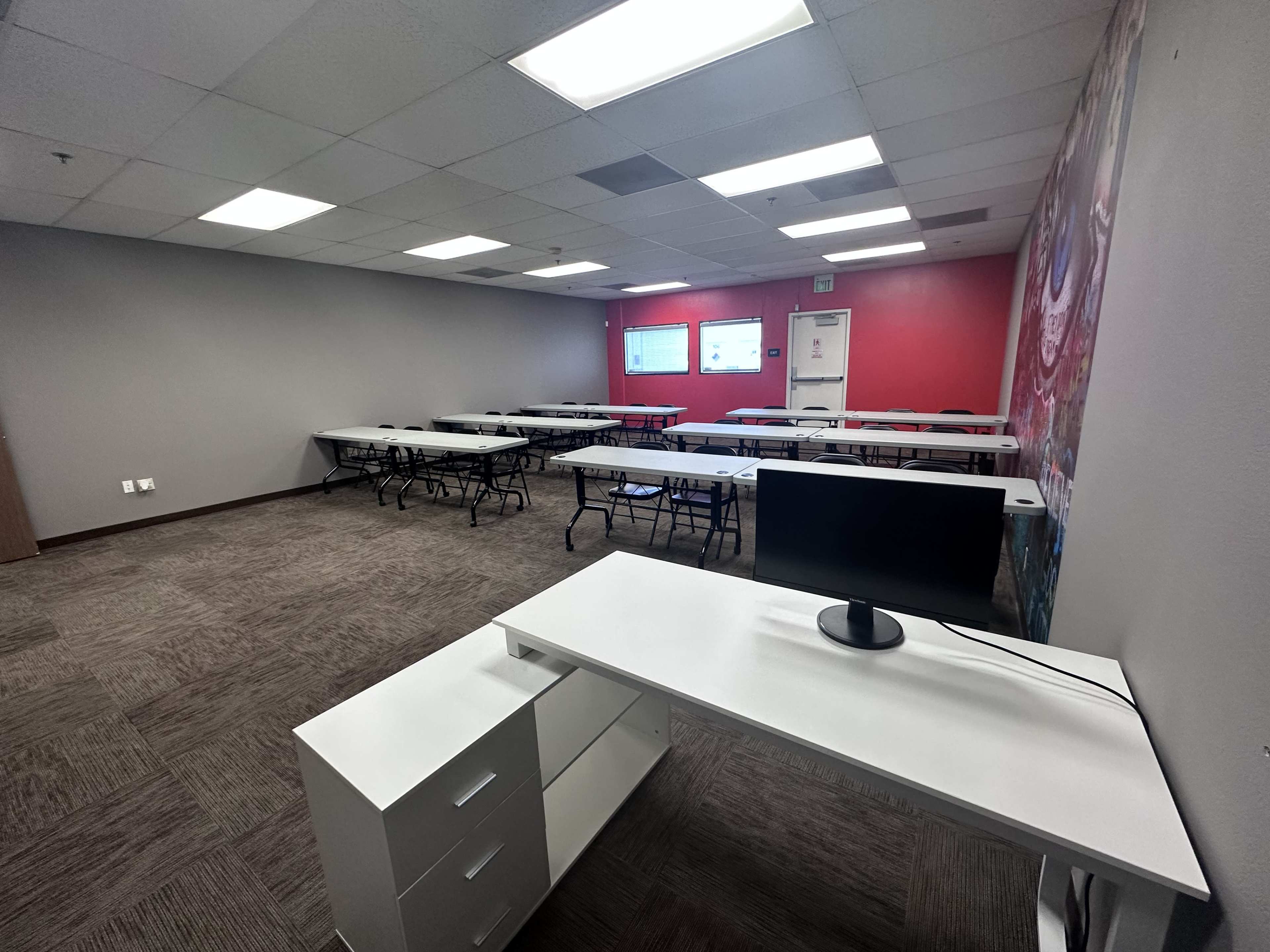A spacious classroom with multiple tables arranged for seating, a white desk with a monitor in the foreground, and a red accent wall.