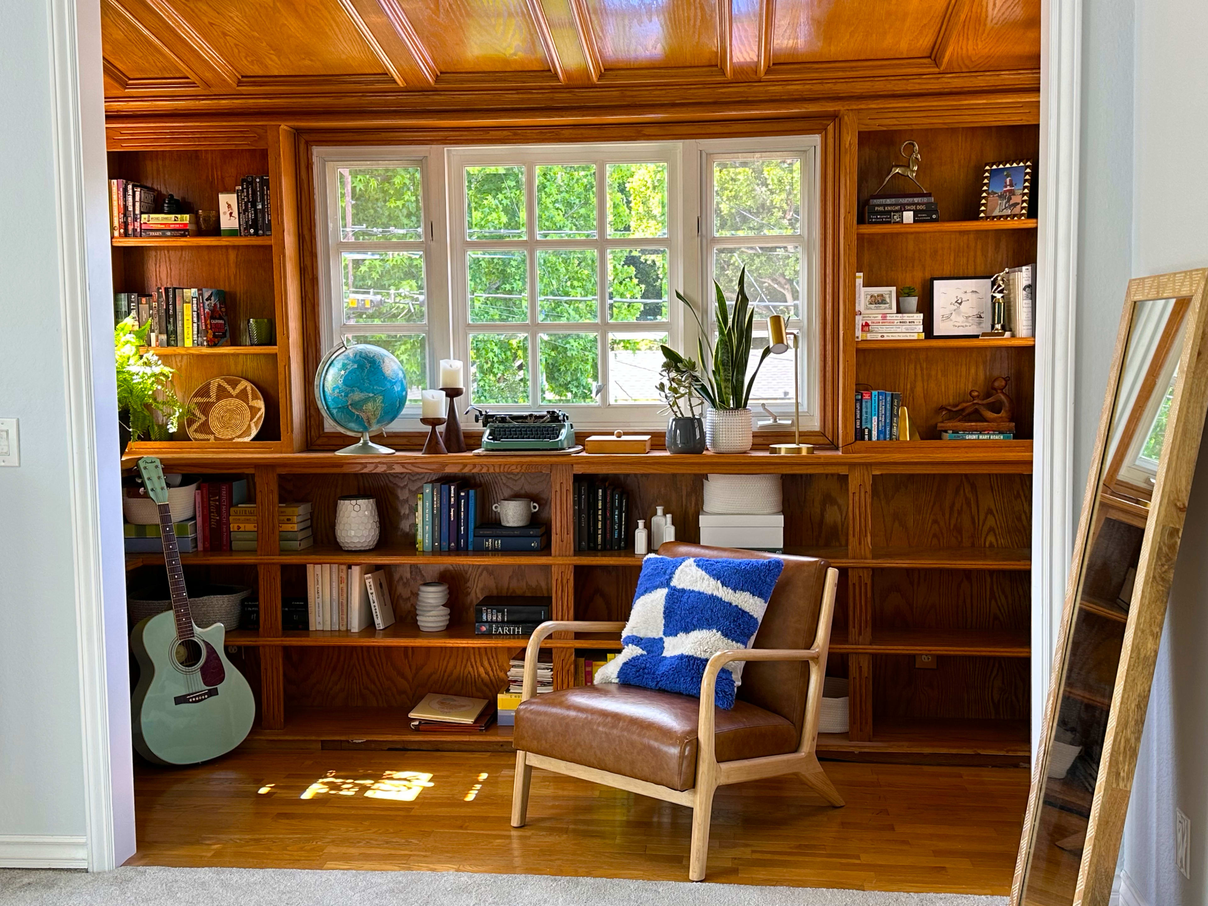 The image shows a wooden bookshelf filled with books and decor, framed by a large window that lets in natural light, accompanied by a comfortable chair and a blue and white throw blanket.