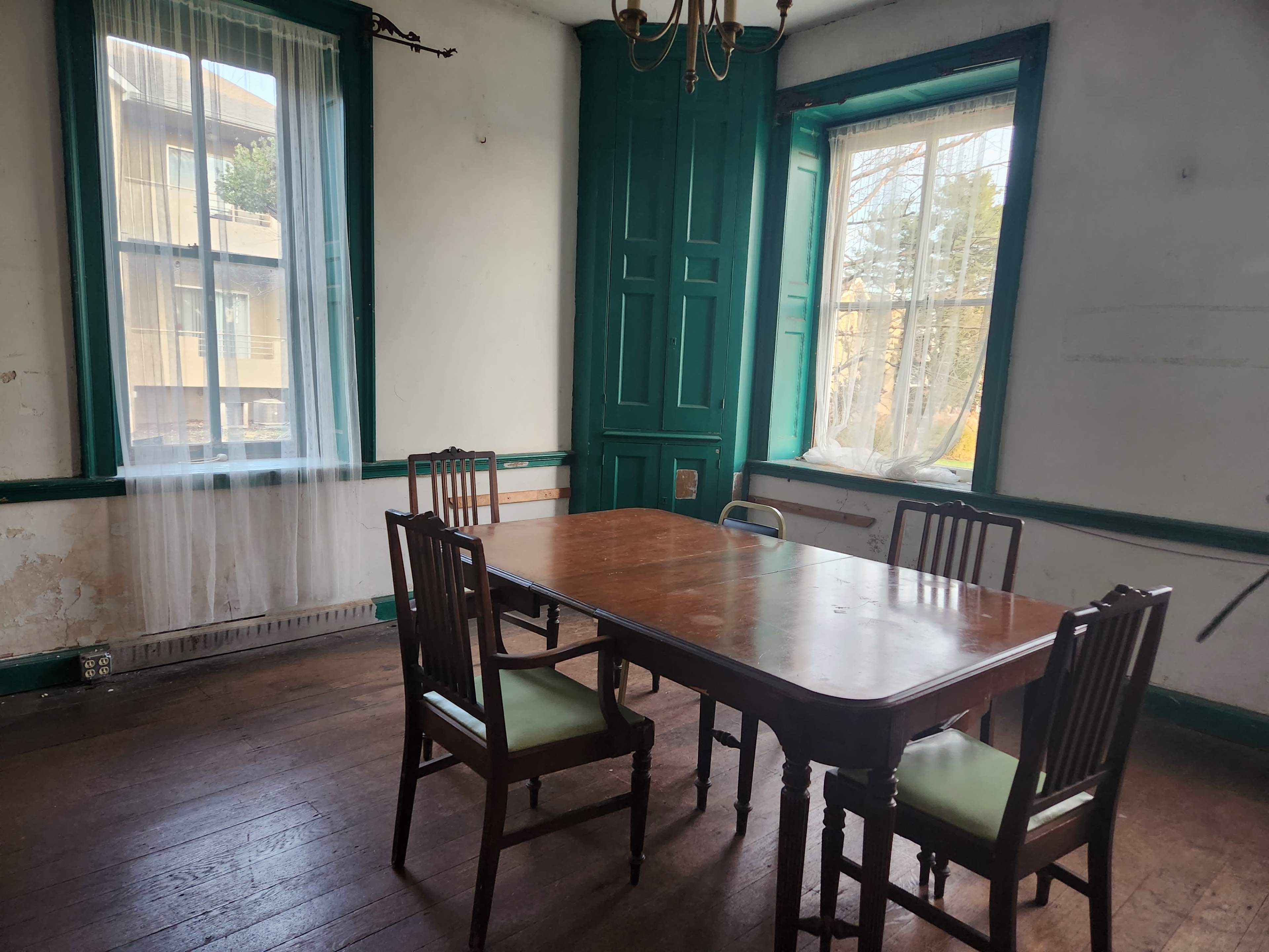 A wooden table surrounded by four chairs is positioned in a room with green accents and large windows.