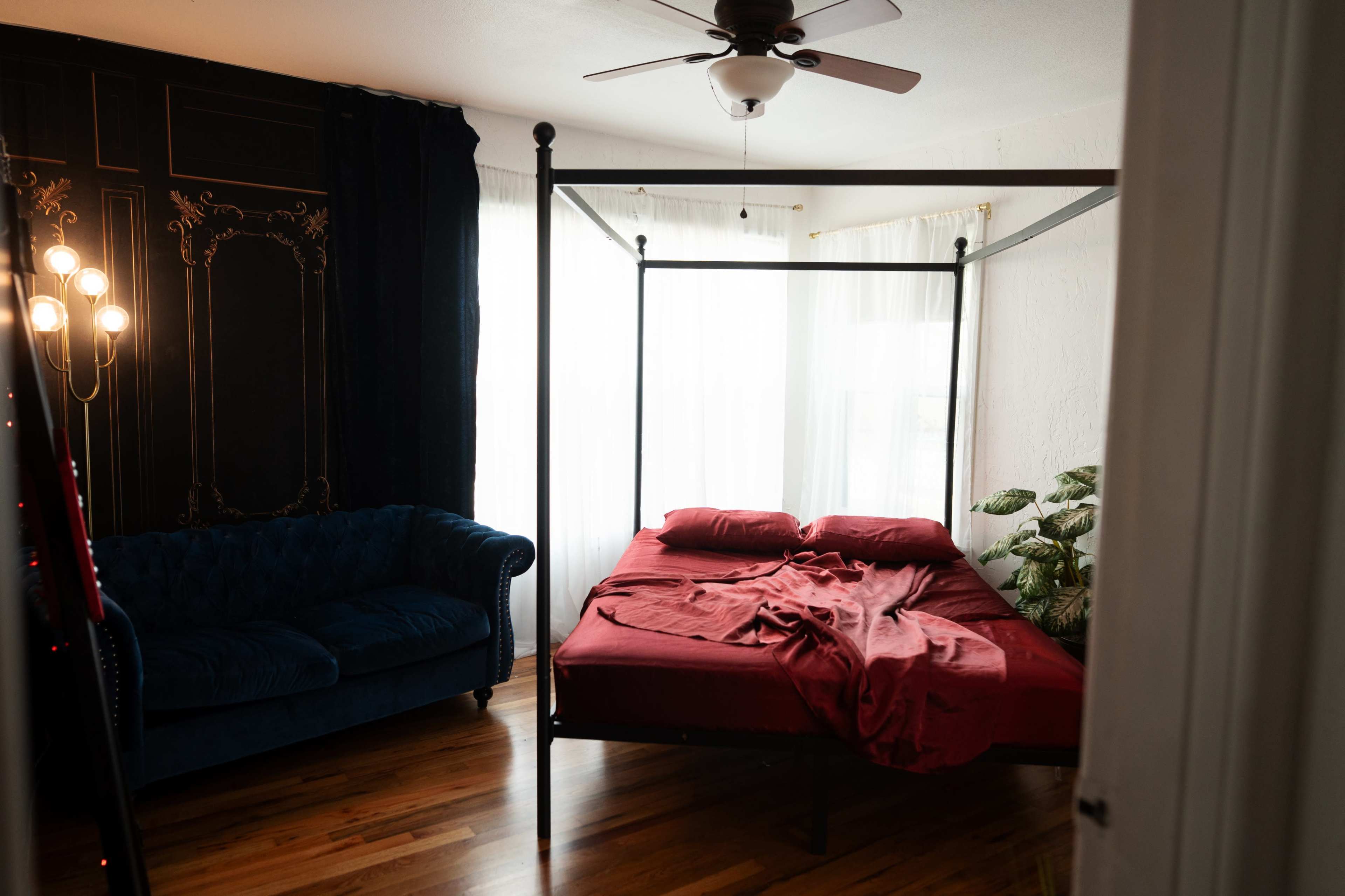 The image shows a bedroom with a four-poster bed made with red sheets and a blue velvet sofa nearby, illuminated by natural light from the windows.