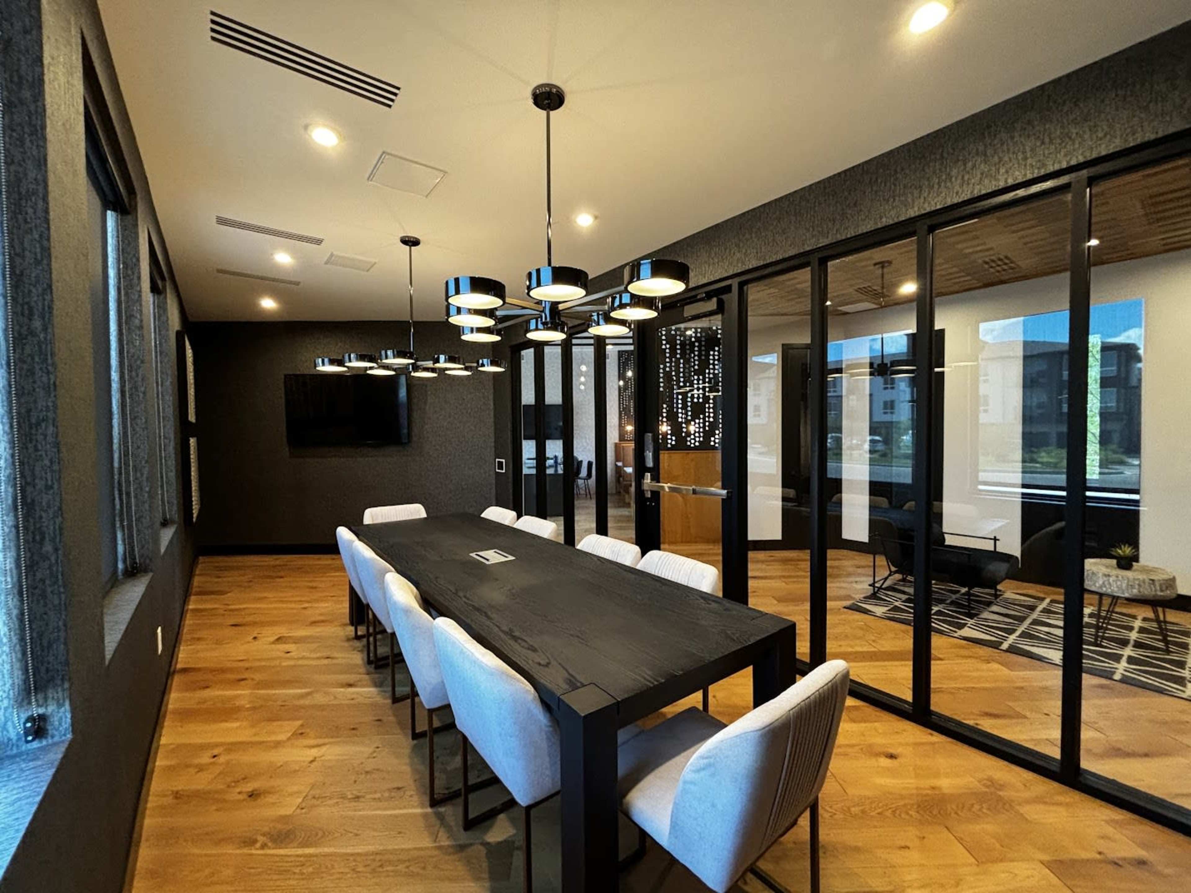 The image shows a modern conference room with a long black table, eight light-colored chairs, and large glass windows, featuring contemporary lighting fixtures and wooden floors.
