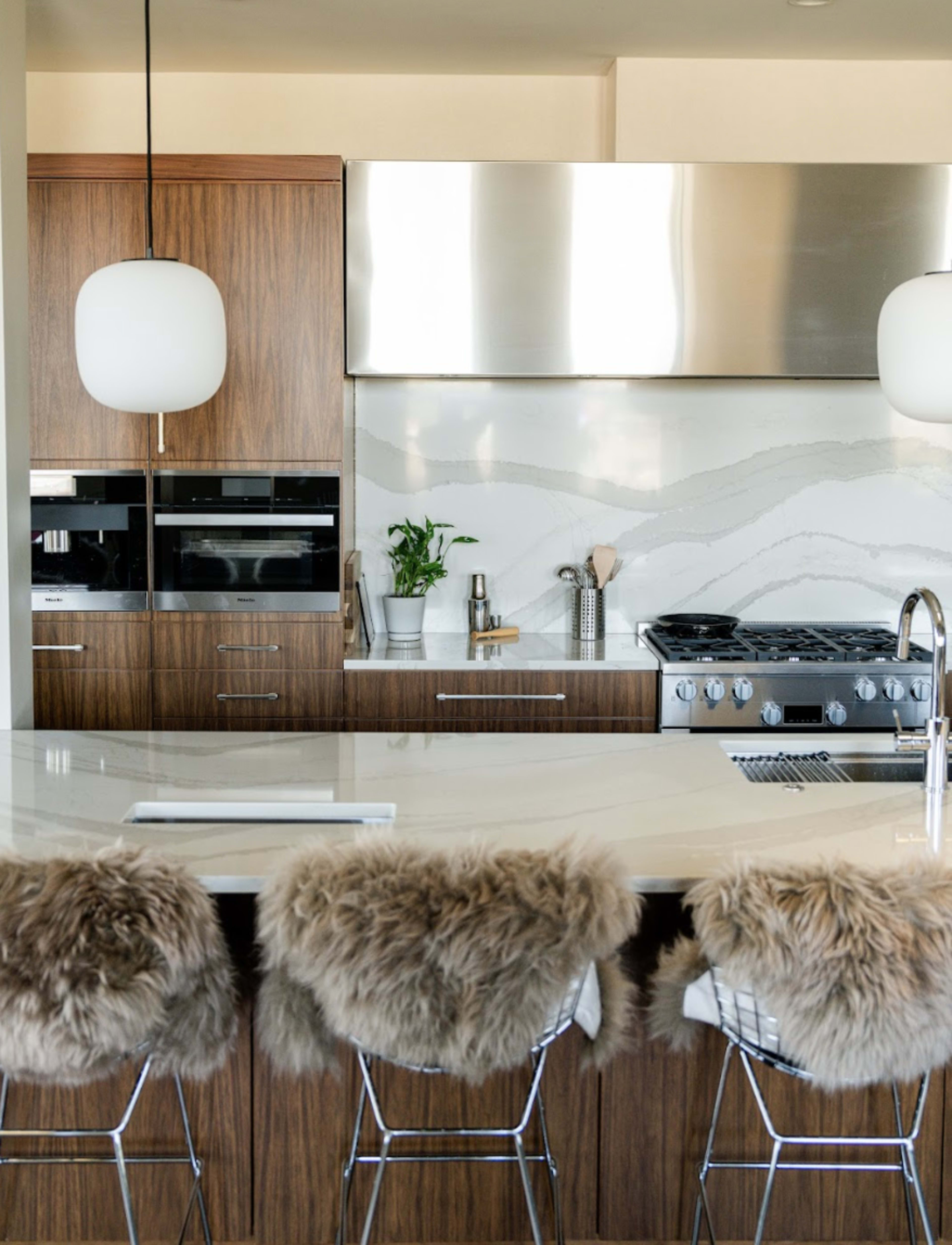 The image showcases a modern kitchen with wooden cabinetry, stainless steel appliances, and barstools with fur seat covers.