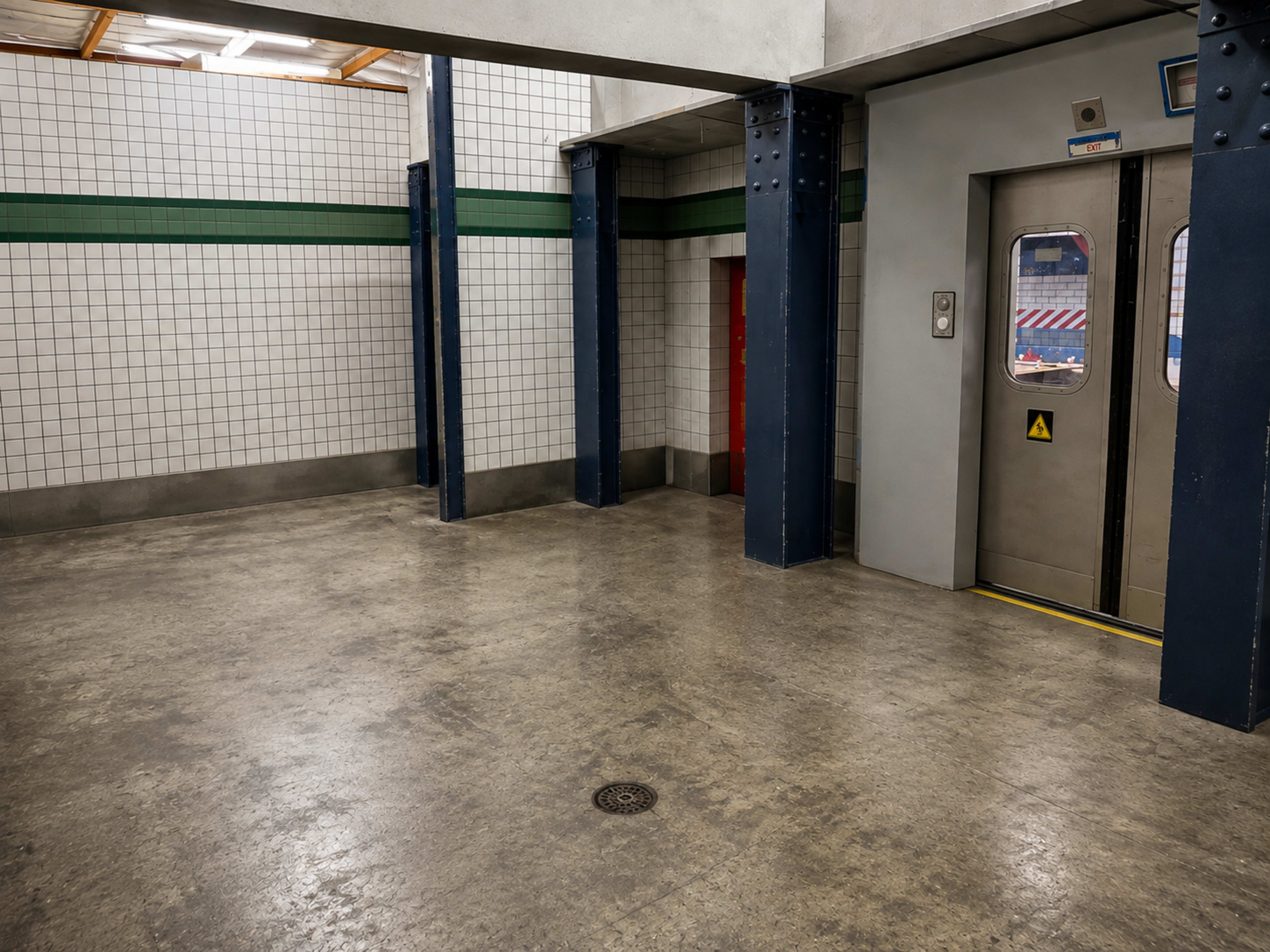 The image shows an empty interior of a subway station with tiled walls, blue pillars, and an elevator door on the right.