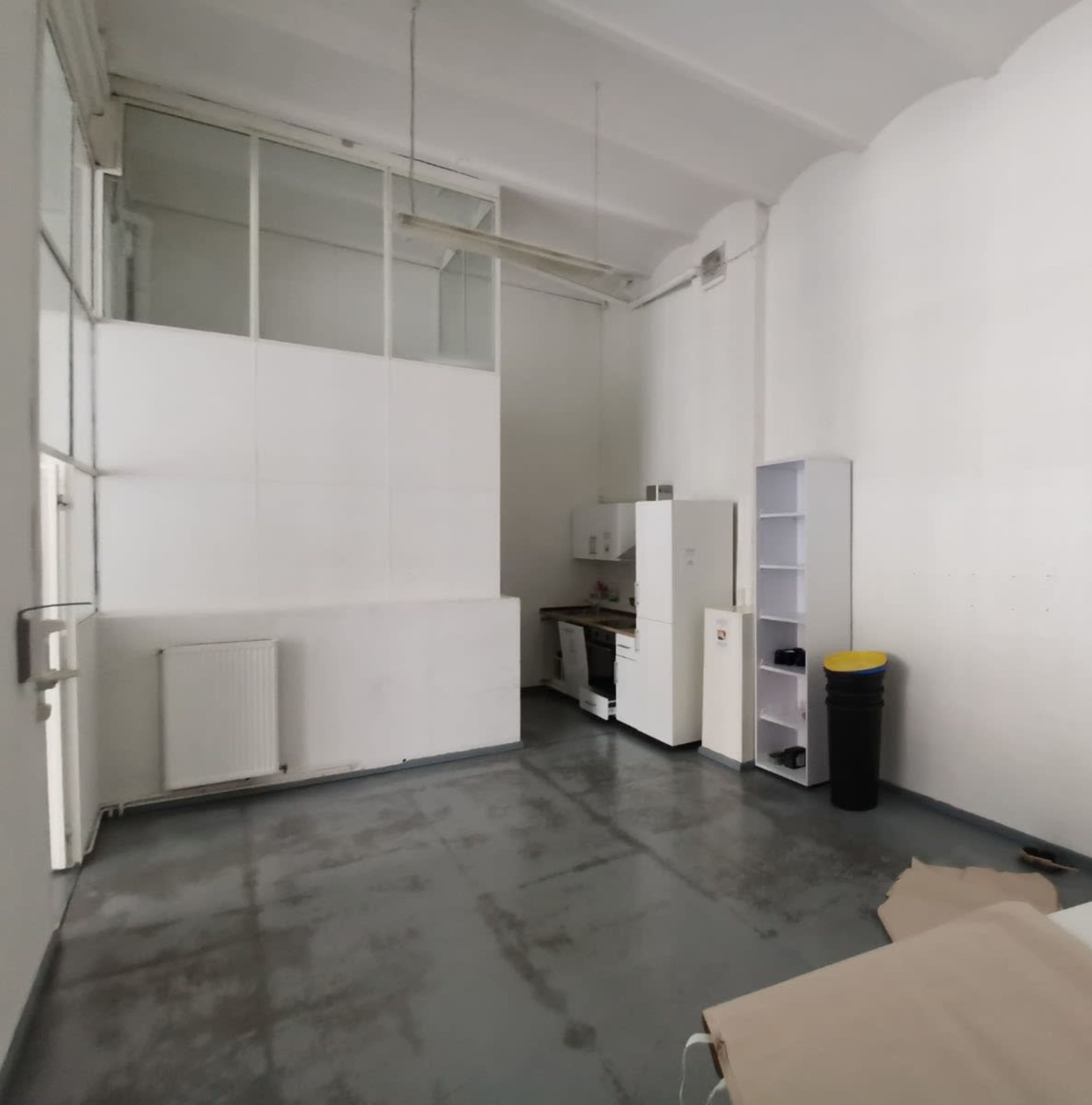 The image shows a minimalist room with gray flooring, white walls, a kitchenette with a refrigerator and stovetop, and a black trash can.
