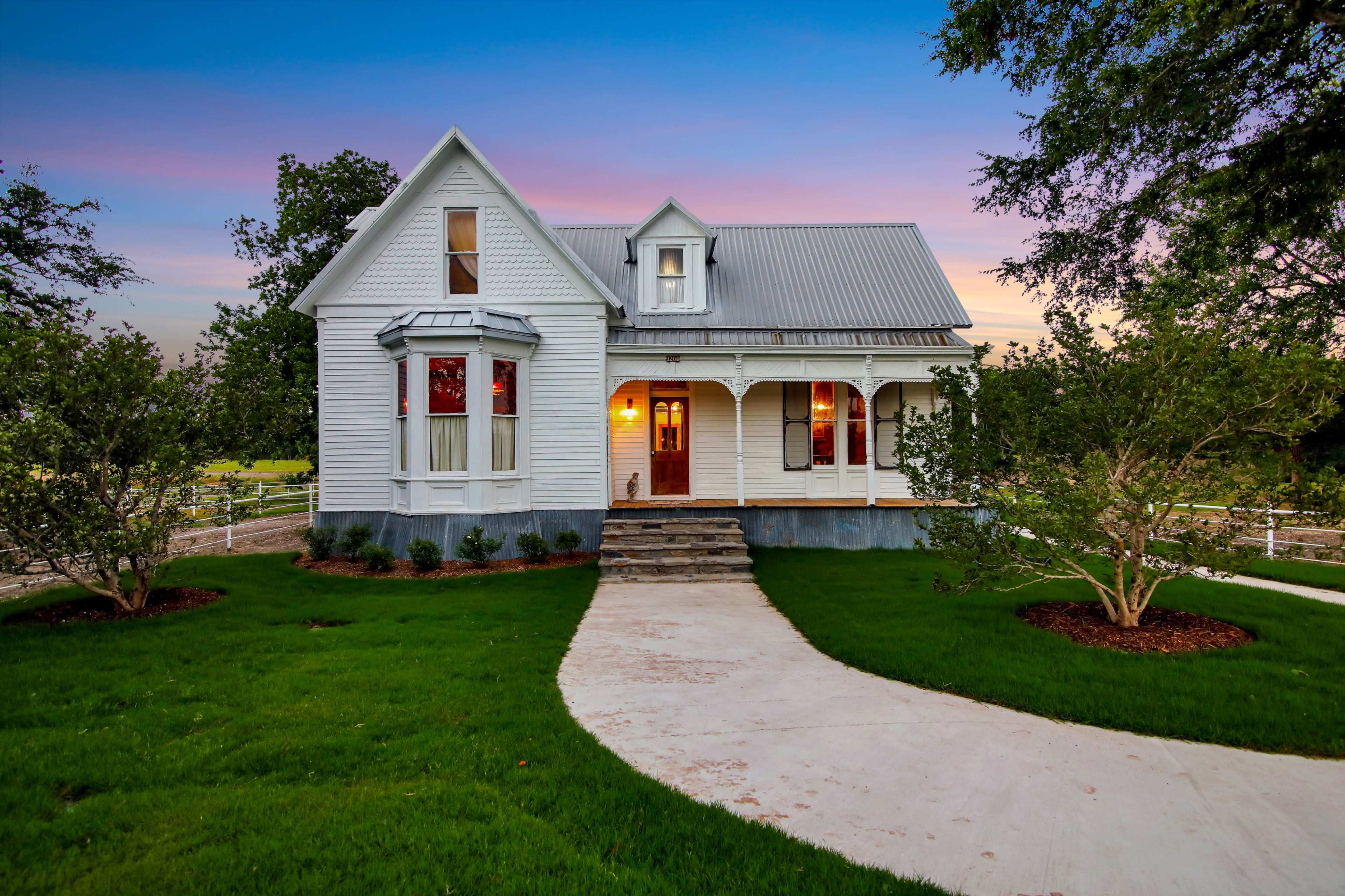A white, two-story Victorian-style house with a metal roof is surrounded by a neatly landscaped lawn and a curved walkway.