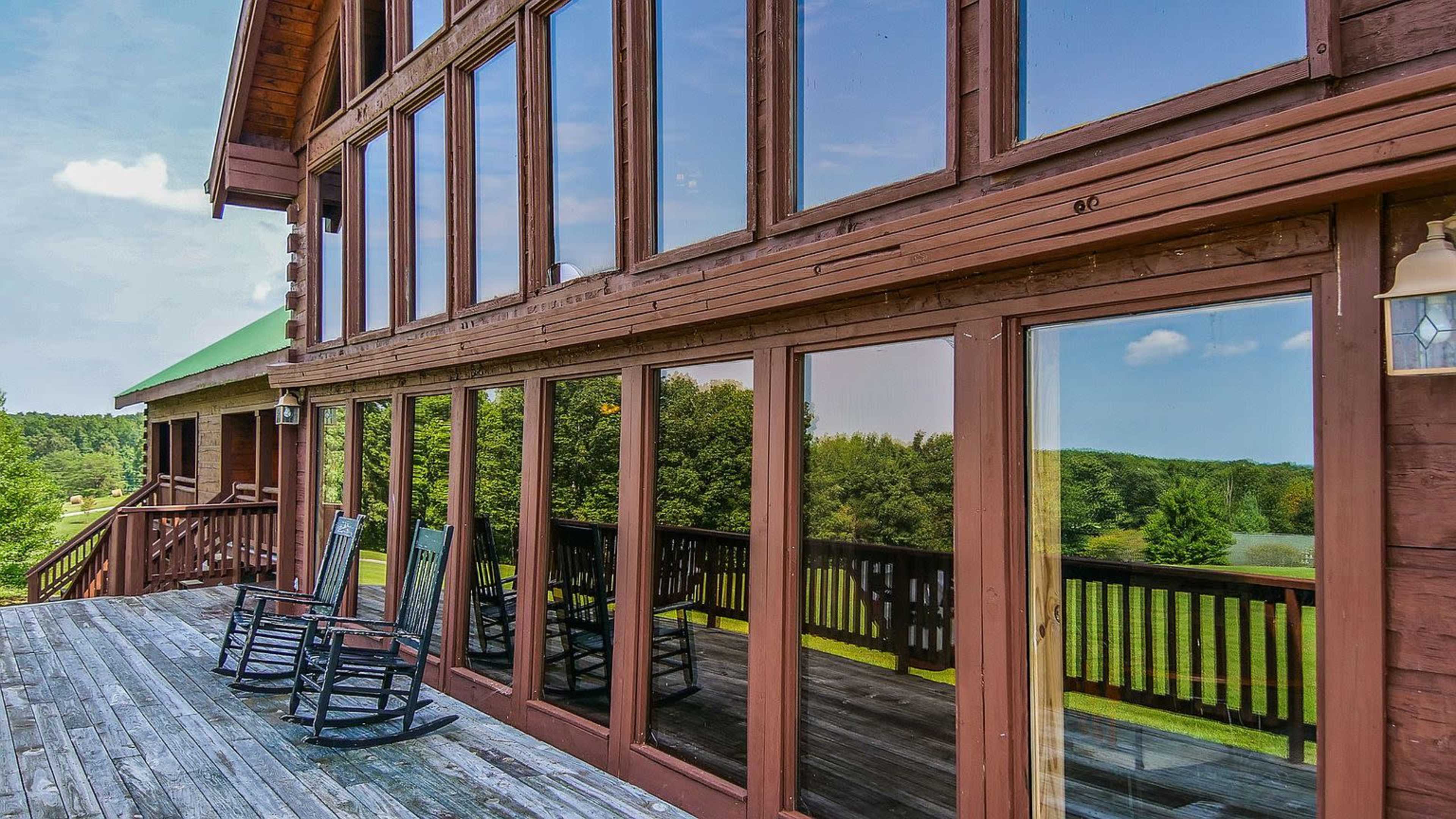 The image shows a wooden deck with rocking chairs in front of a large wall of windows reflecting a green landscape.