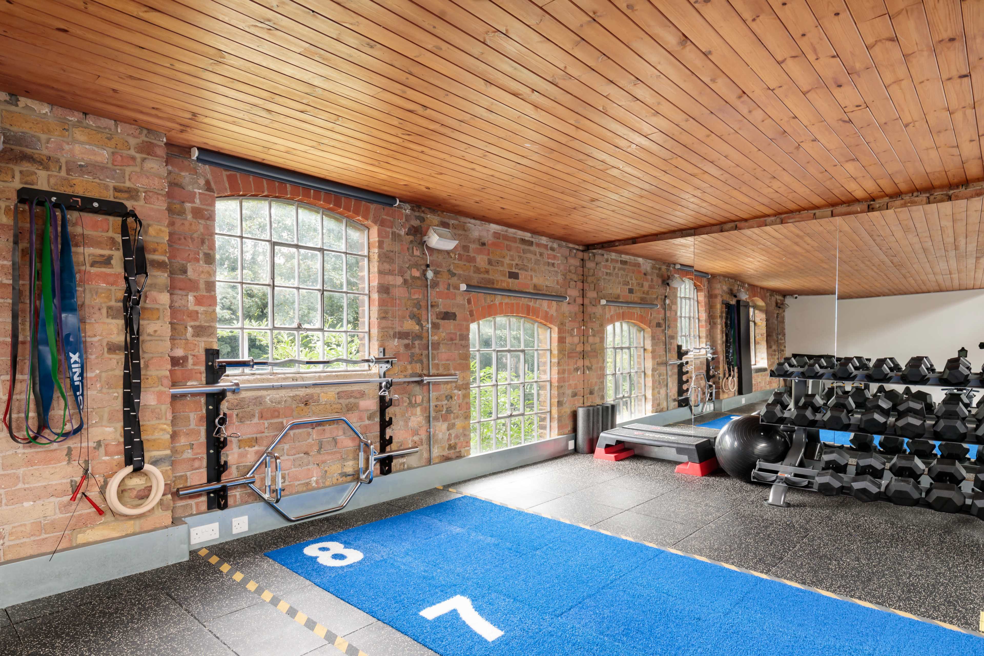 A spacious gym with brick walls, large windows, a blue exercise mat, and various exercise equipment including dumbbells and rings.