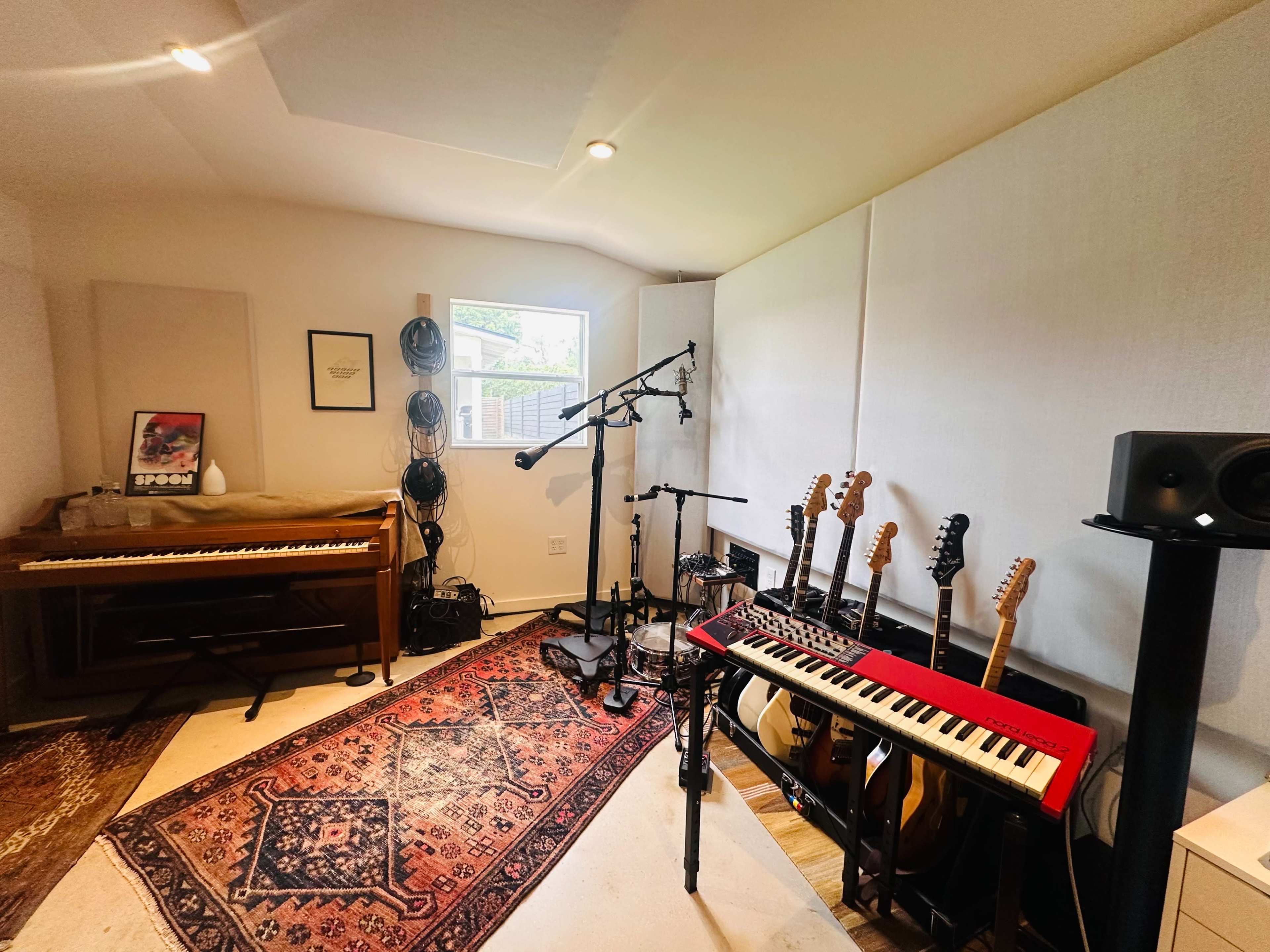 The image shows a recording studio with a piano, several guitars, a microphone setup, and an electronic keyboard on a stand, all arranged on a patterned rug.