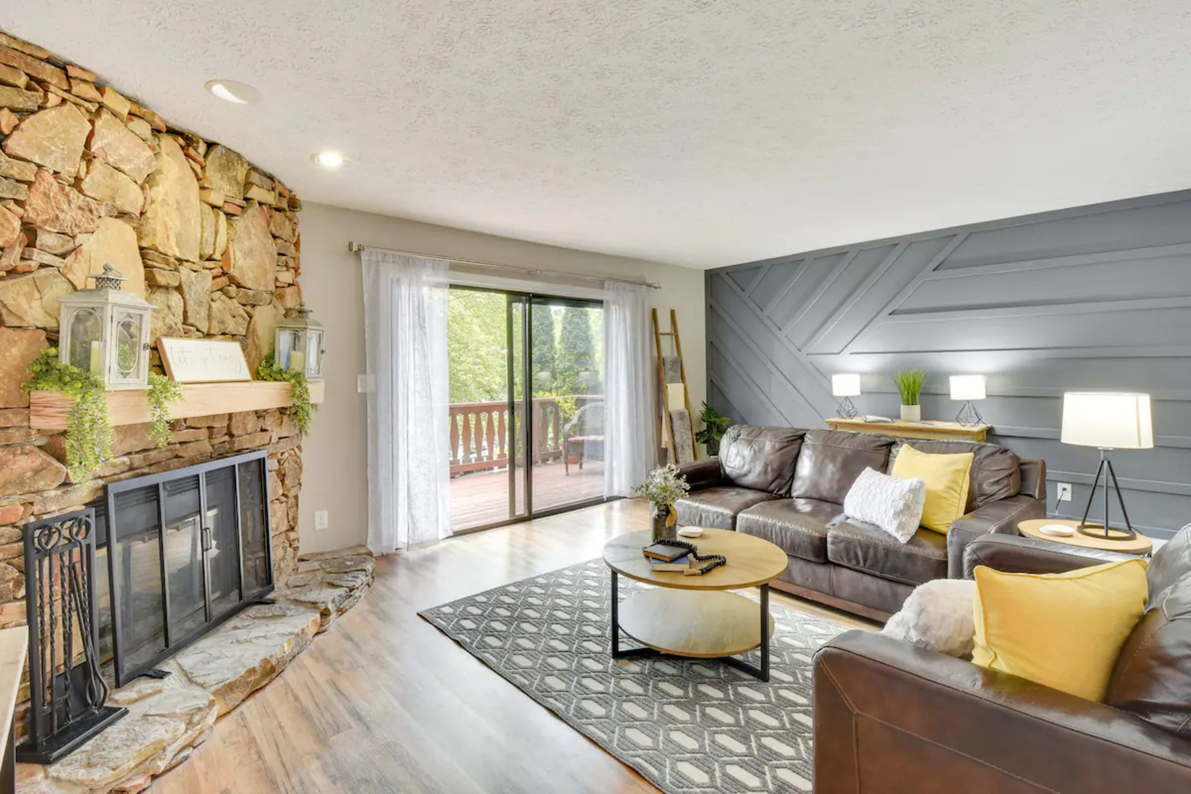 A living room with a stone fireplace, a large window leading to a wooden deck, and a mix of dark leather furniture and decorative accents.