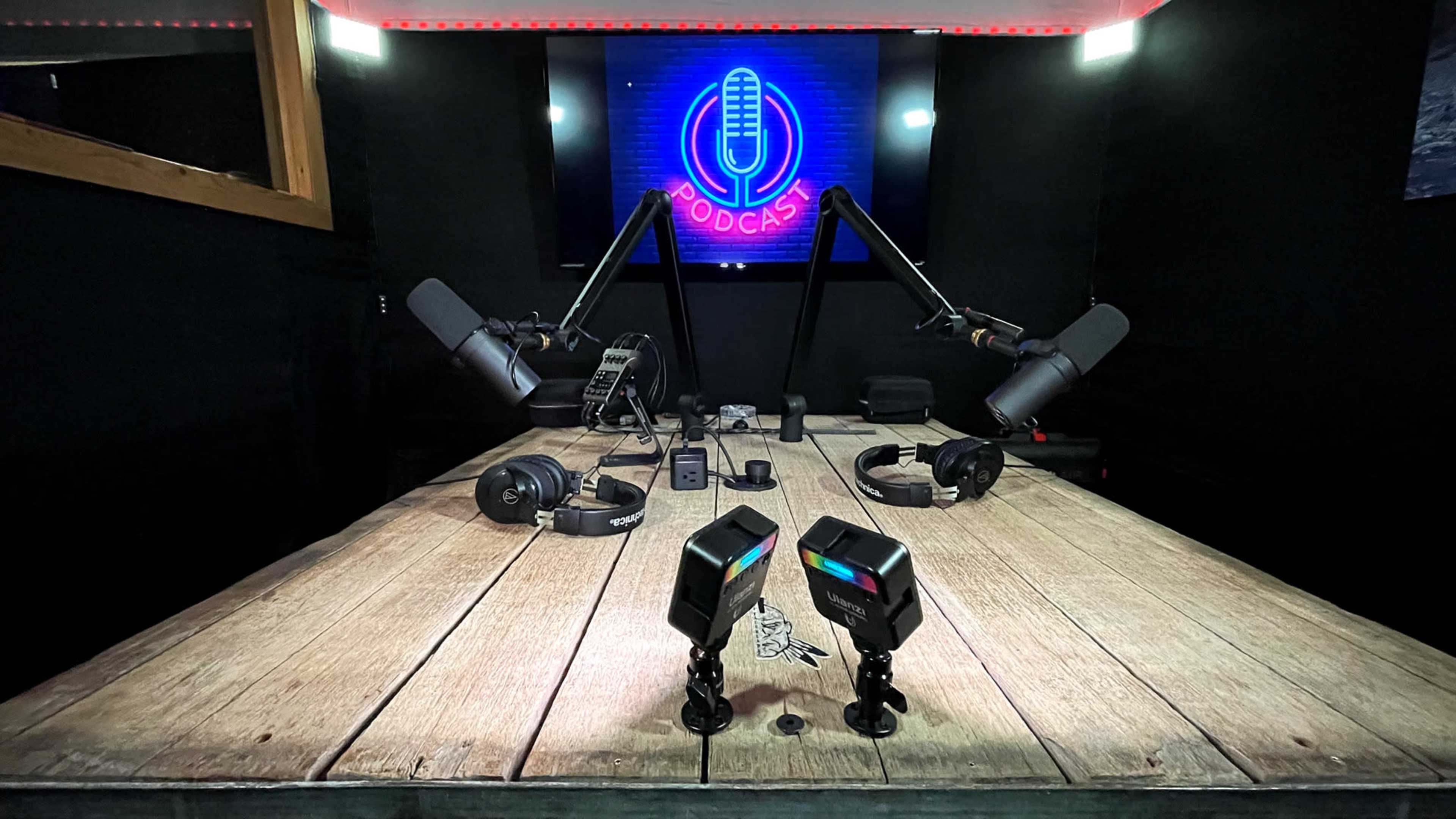 A podcasting studio with a wooden table, microphones mounted on adjustable arms, headphones, and a screen displaying a microphone logo.