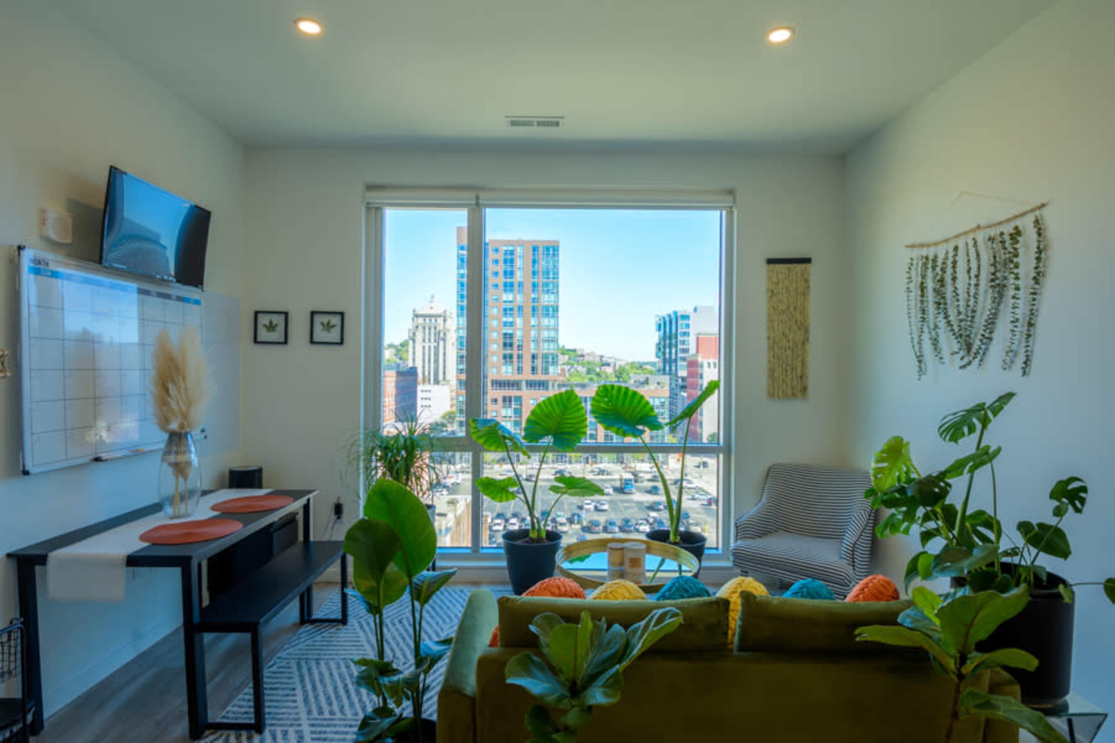A modern living room features a large window with a city view, colorful cushions on a couch, and various indoor plants.