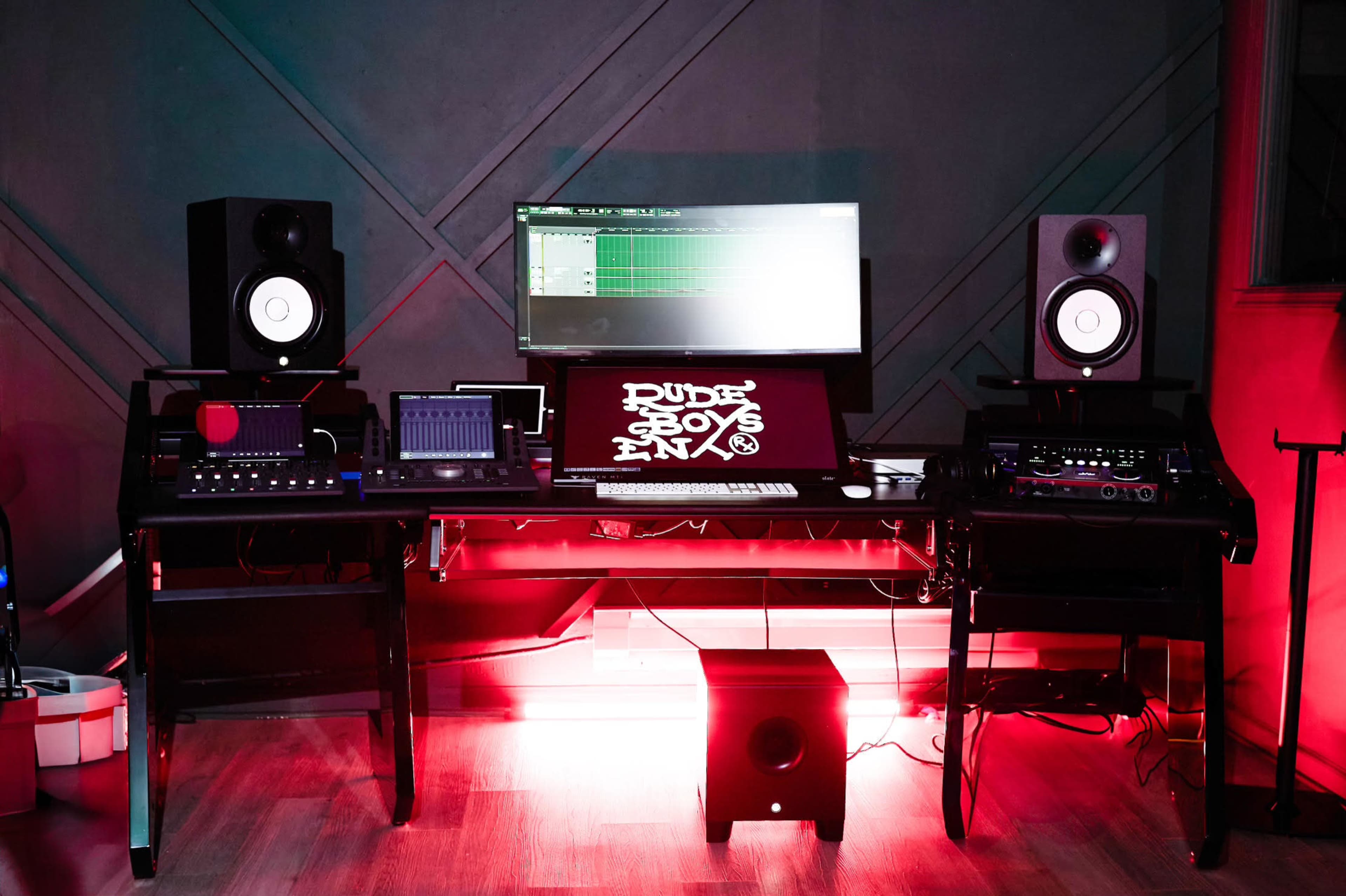 A music production studio setup featuring a desk with various audio equipment, monitors, and a large screen displaying a digital audio workstation.