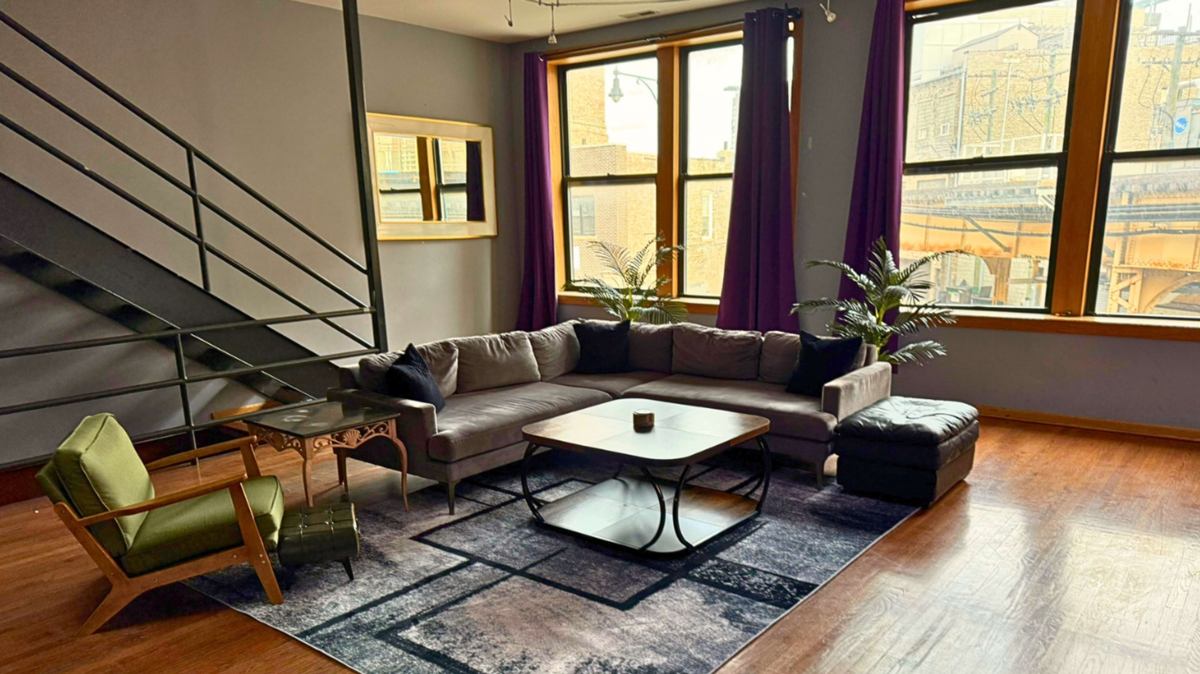 A modern living room features a sectional sofa, a coffee table, and a green armchair, with large windows revealing an urban view.