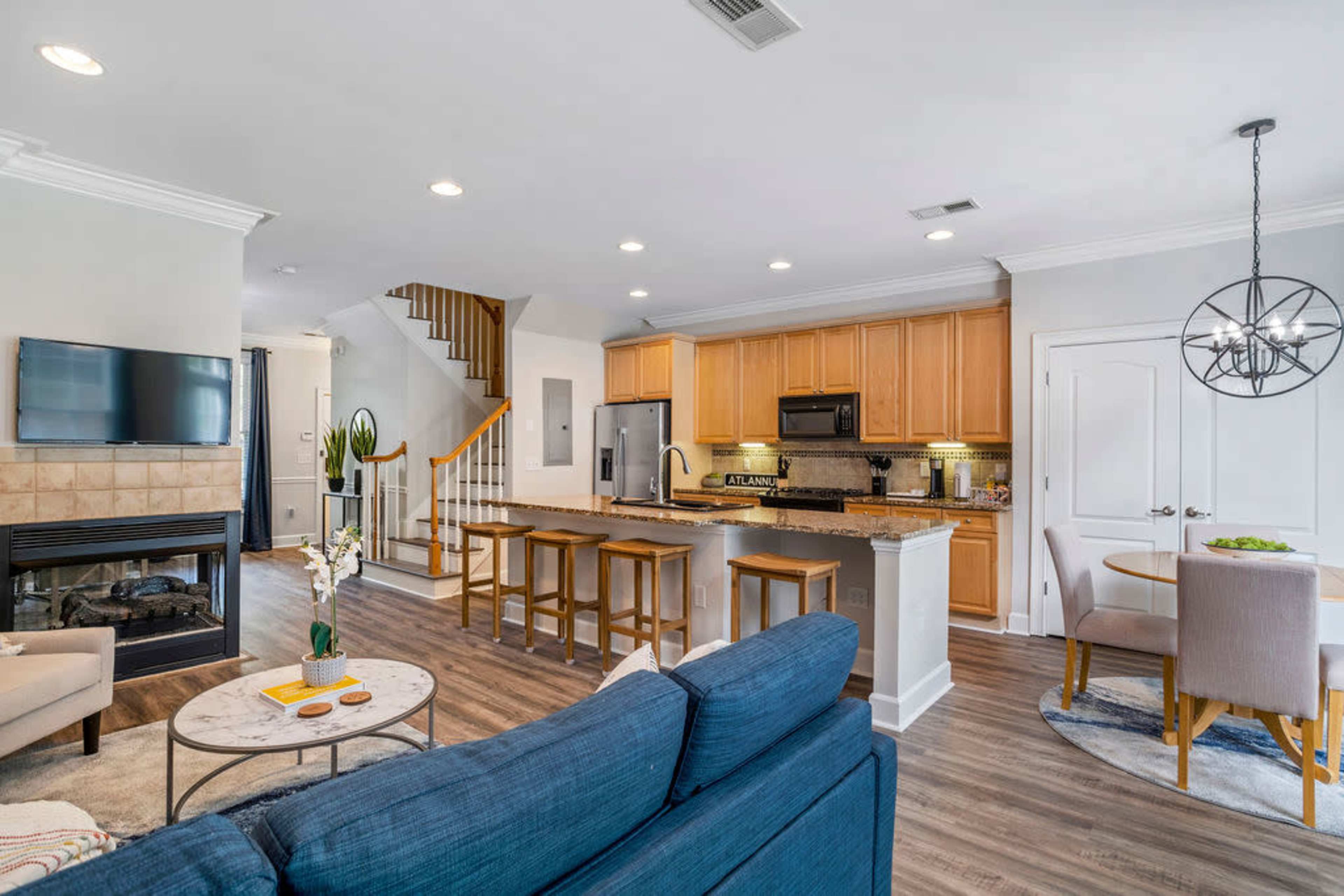 The image shows a modern living space featuring a kitchen with wooden cabinets, a dining area, a living room with a blue sofa, and a staircase leading to an upper level.