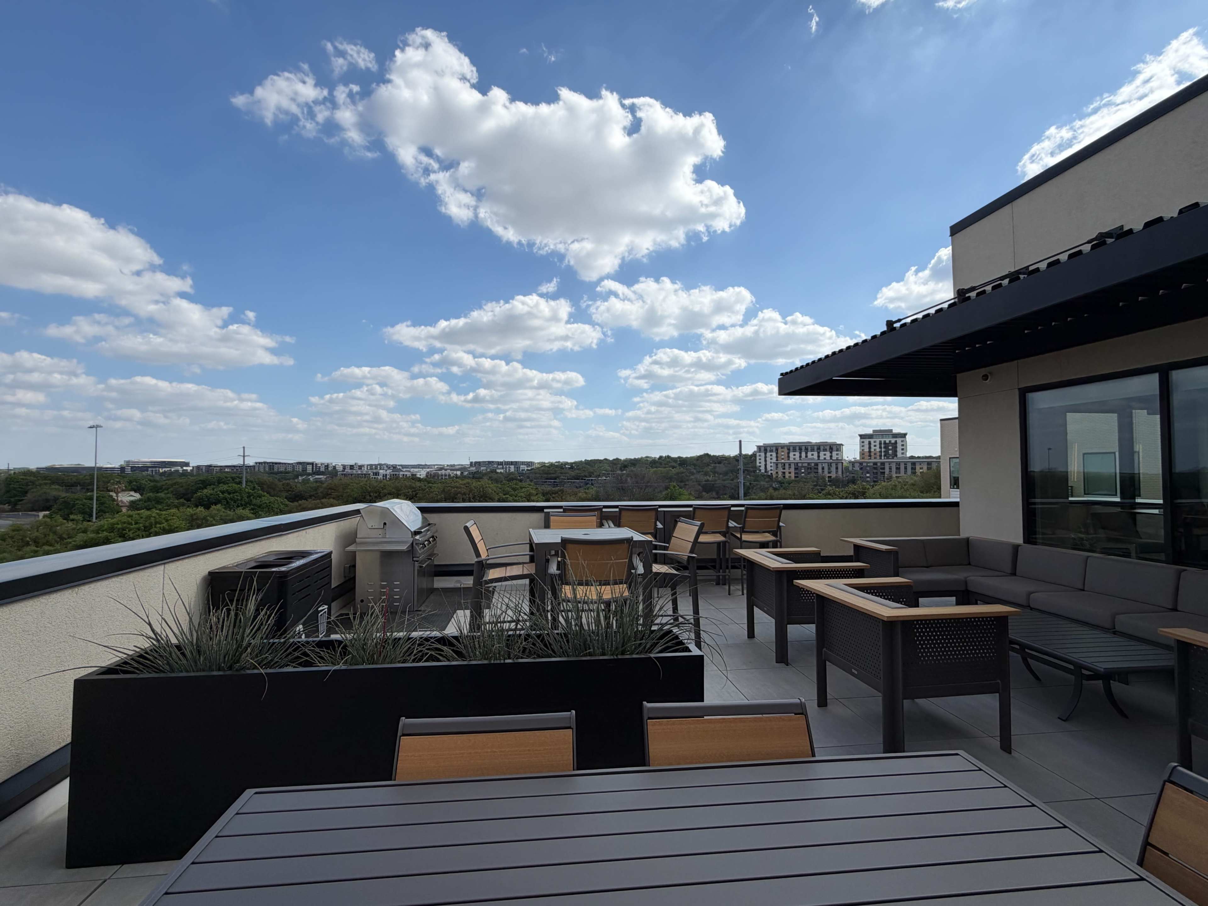 A rooftop patio features wooden tables and chairs amidst green landscaping, with a clear blue sky and scattered clouds in the background.