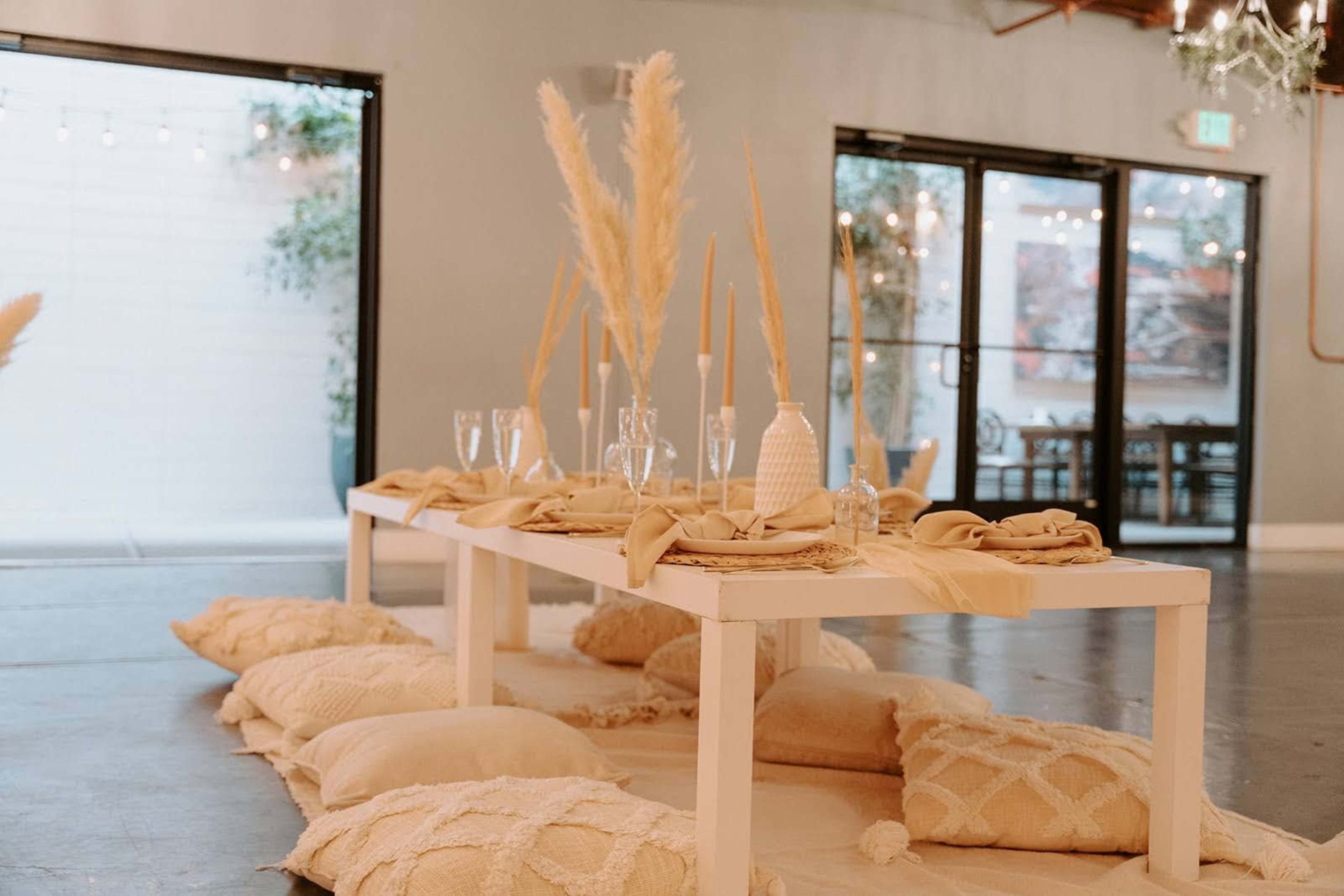 A low white table is set with simple tableware and decorated with pampas grass, surrounded by floor cushions in a softly lit indoor space.
