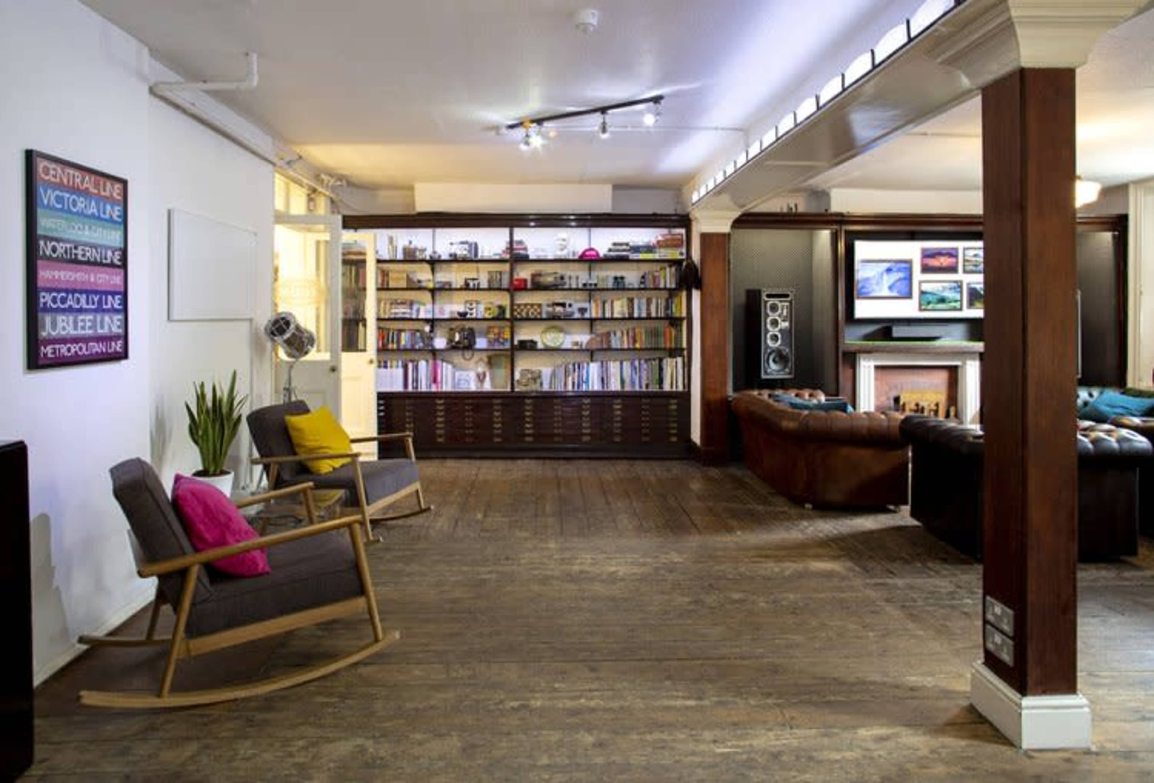 The image shows a spacious interior with wooden flooring, a large bookshelf filled with books and decor items, and a cozy seating area featuring a brown leather couch and a rocking chair.