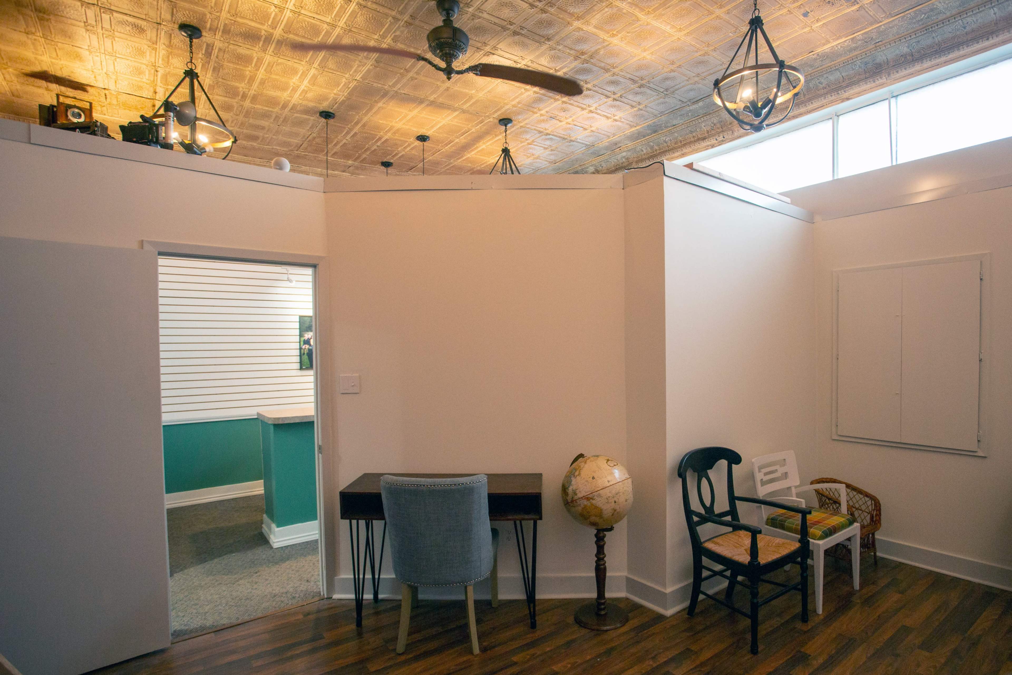 A room with a desk, a globe, and two chairs, leading into a space with a green wall and overhead lights.