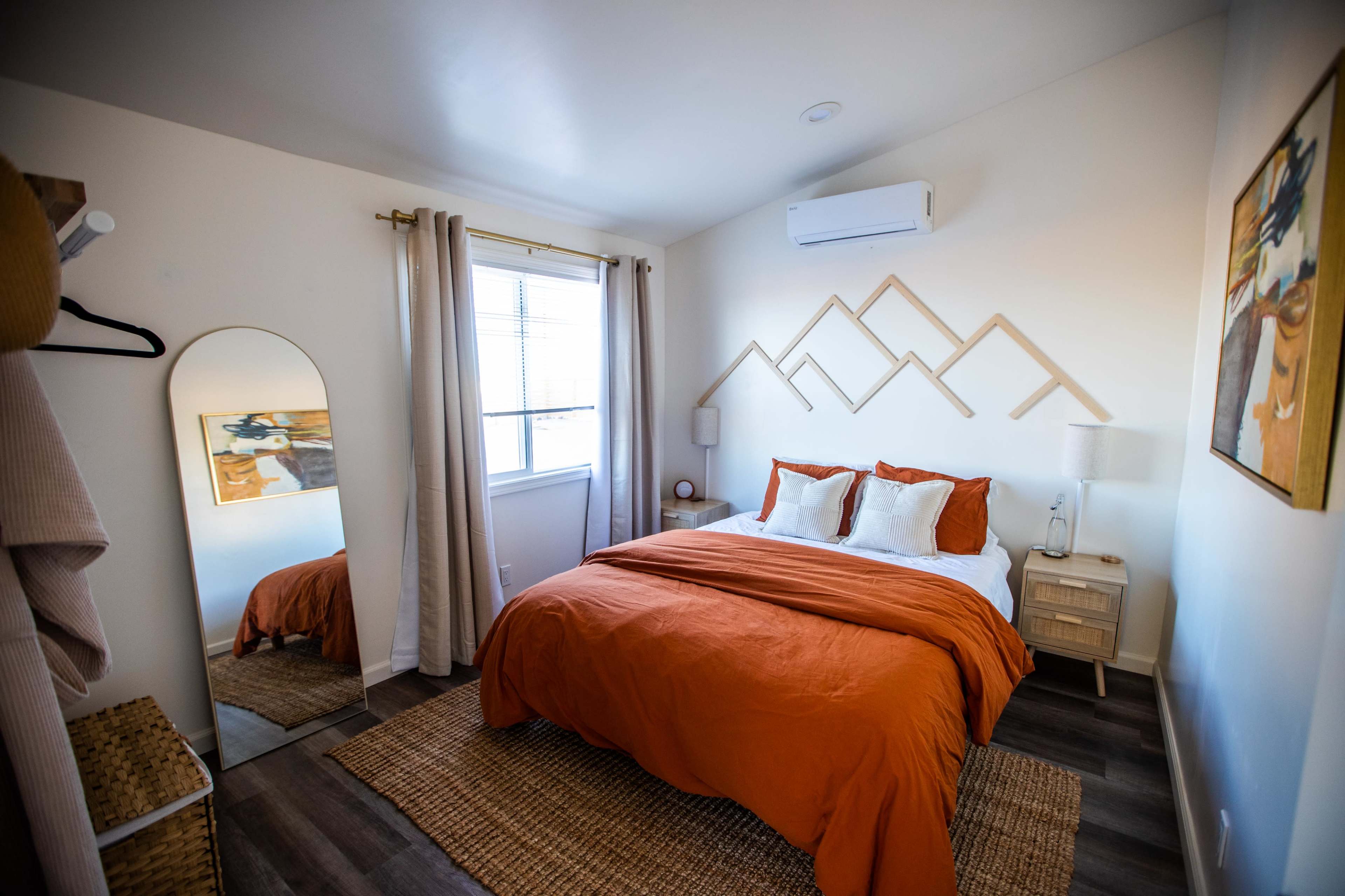 A cozy bedroom with an orange bedspread, a large mirror, and minimalist decor featuring mountain shapes on the wall.