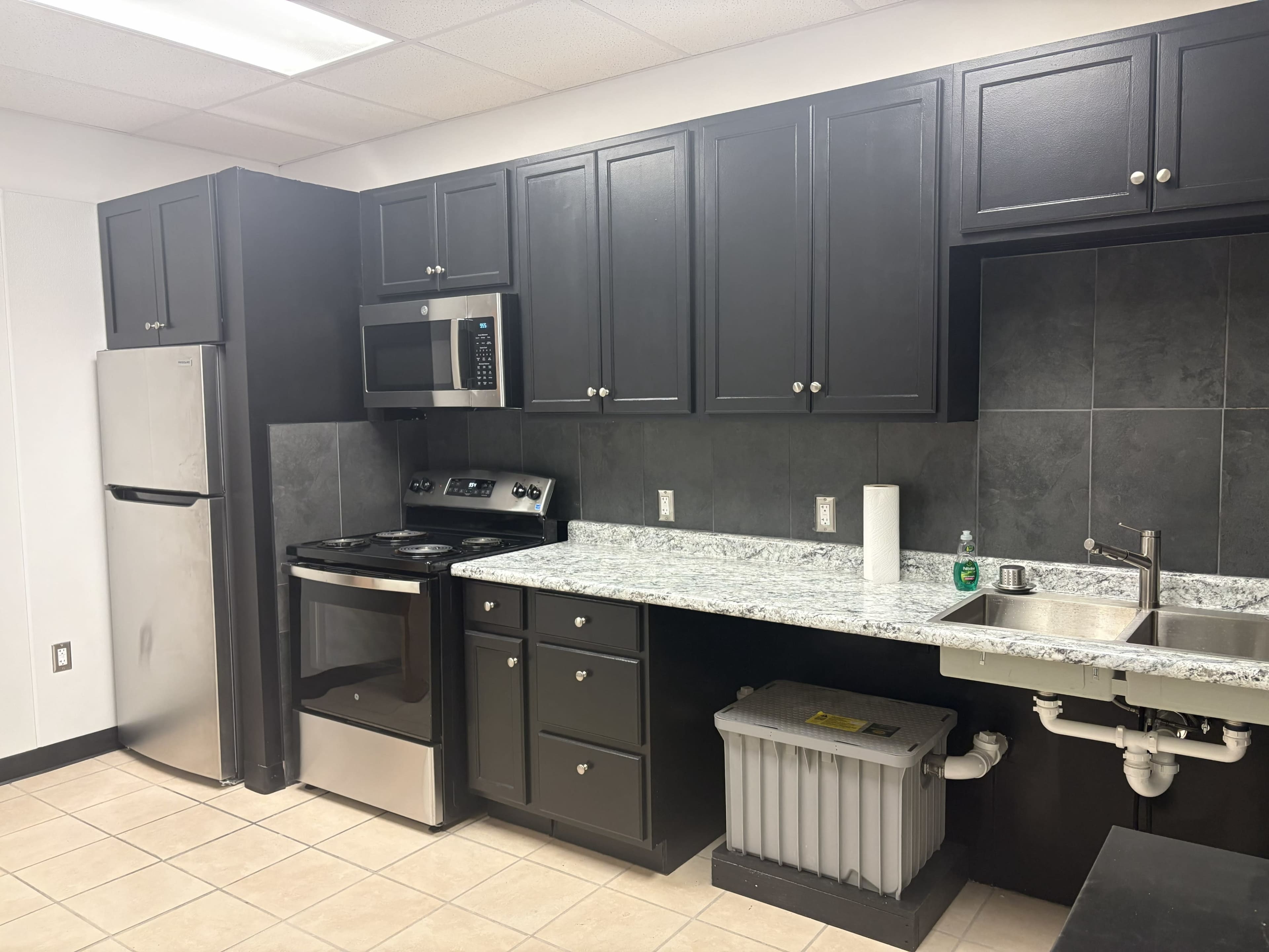 The image shows a modern kitchen featuring black cabinets, a gray countertop, a stainless steel refrigerator, an oven, and a sink with a drainage system visible below.
