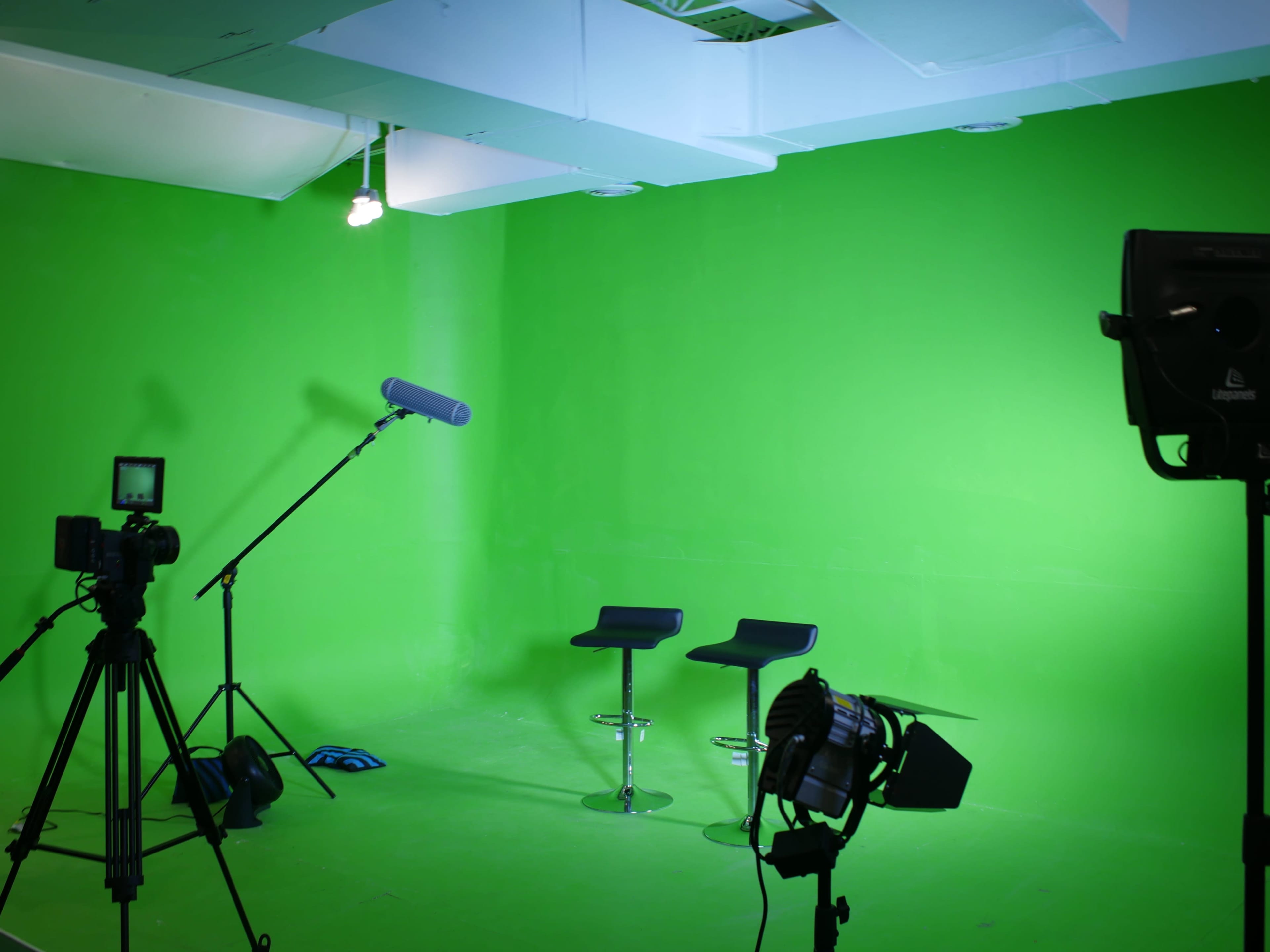 A green screen studio setup with two stools, multiple cameras, a microphone on a boom arm, and lighting equipment.