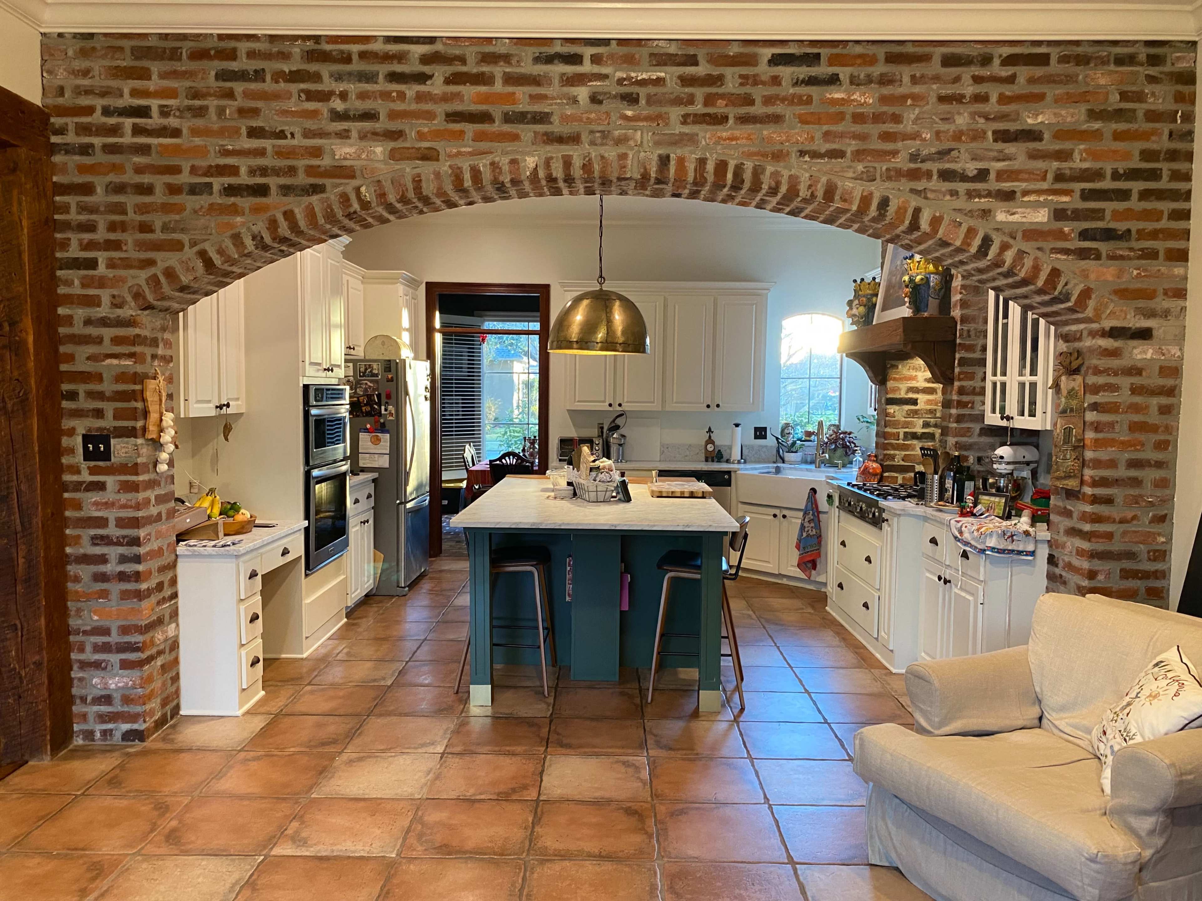 A kitchen with a brick archway, featuring white cabinetry, a large island with bar stools, and tiled flooring.