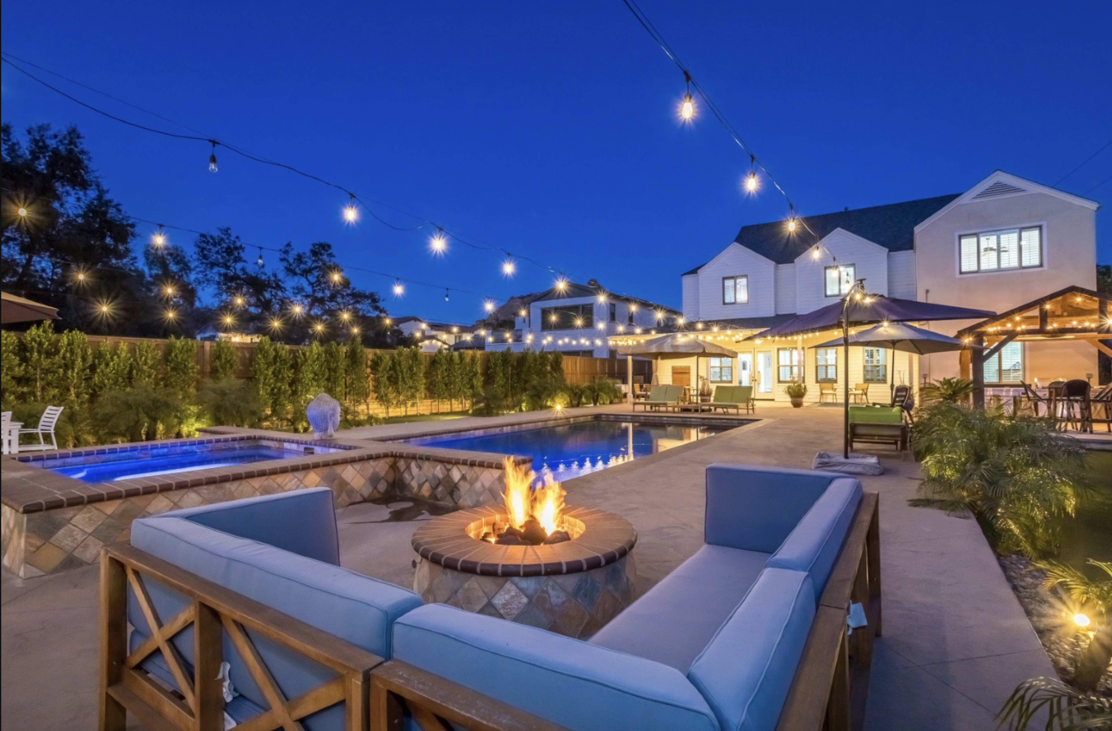 A modern backyard features a fire pit surrounded by seating, a swimming pool, a hot tub, and string lights illuminating the area at night.