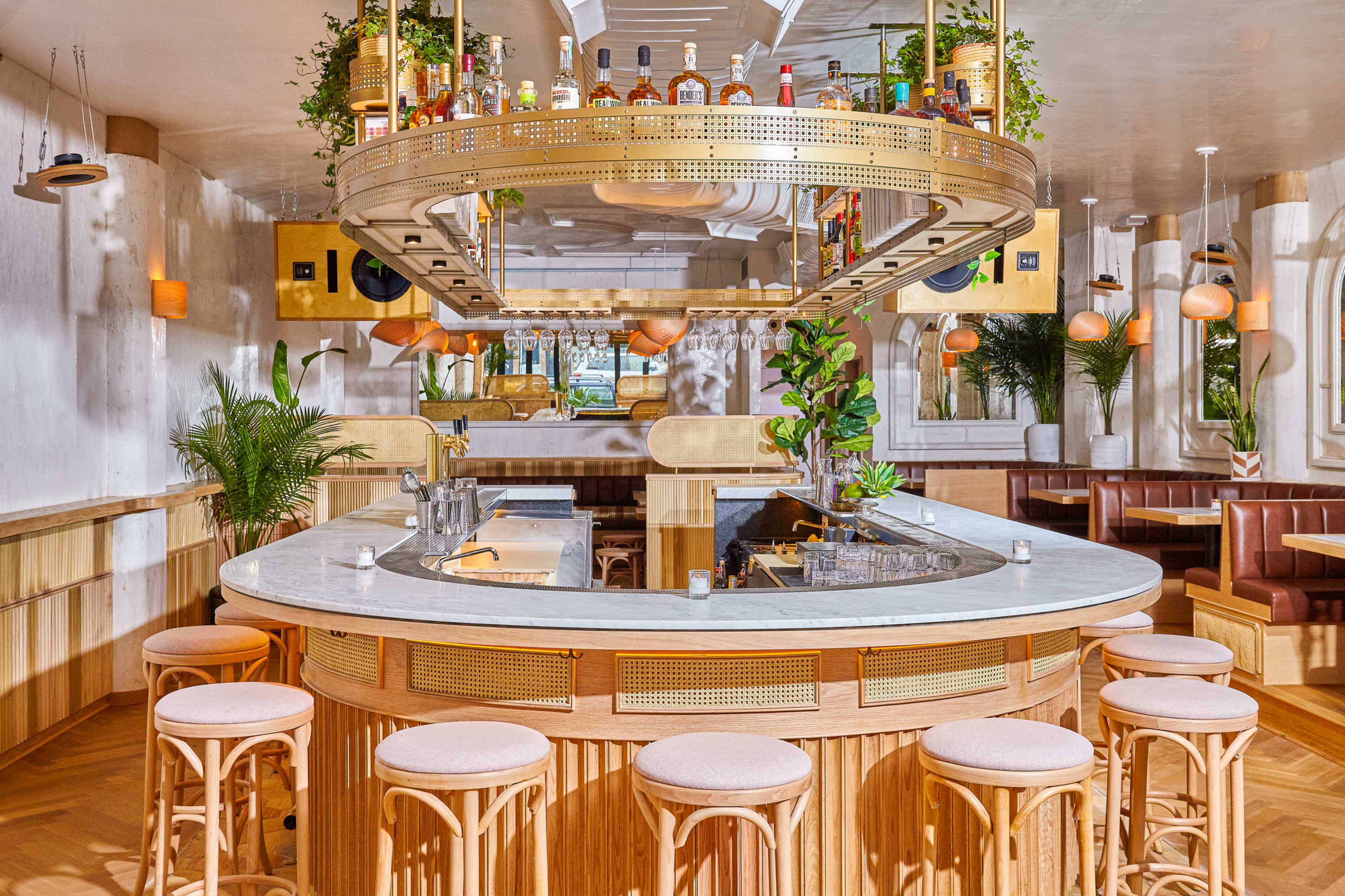 A modern bar area with a circular marble countertop, wooden stools, and decorative plants, featuring shelves stocked with various bottles.