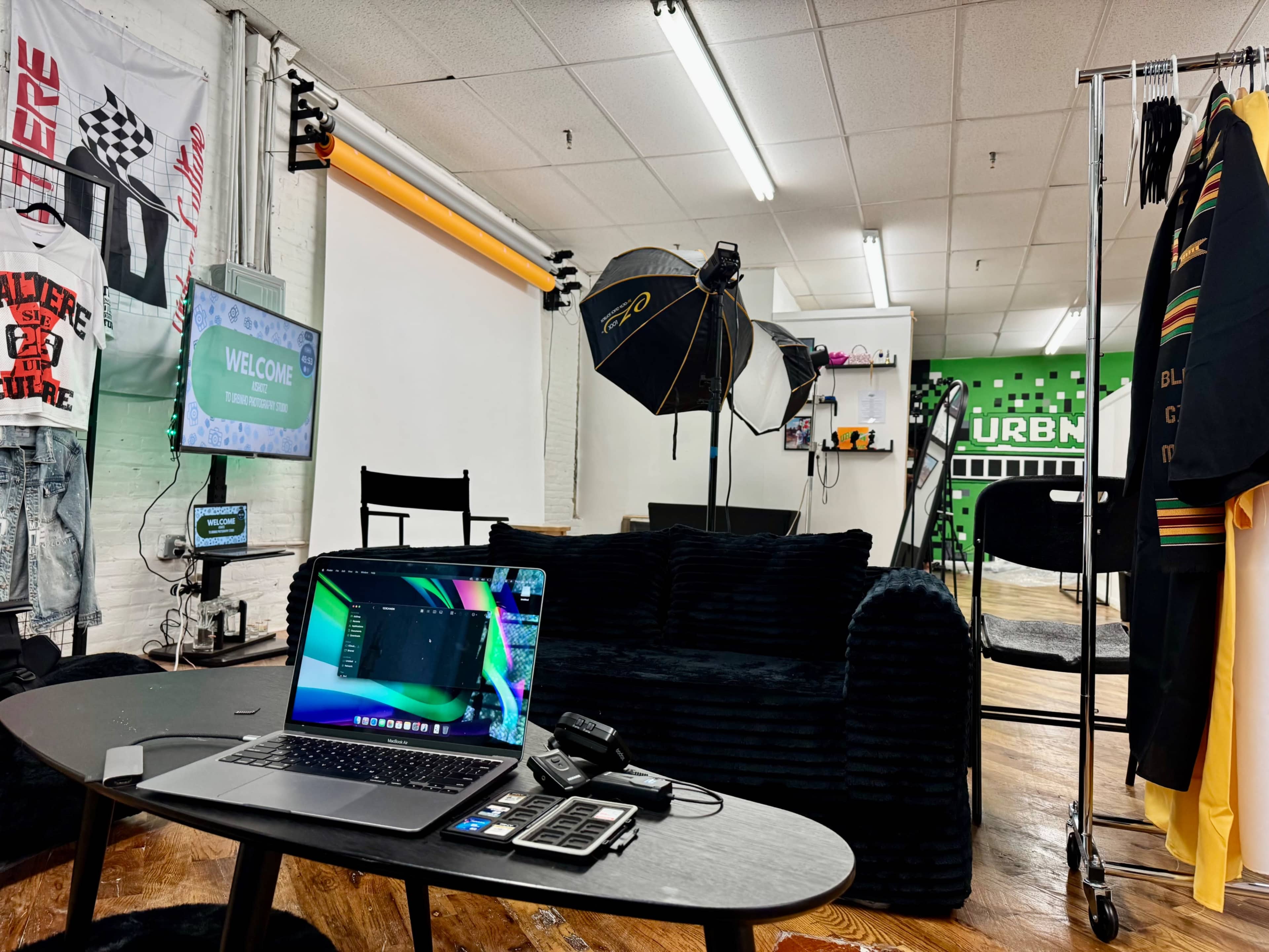 The image shows a well-equipped creative studio featuring a laptop on a table, a black couch, clothing displays, and photography equipment against a vibrant green wall.