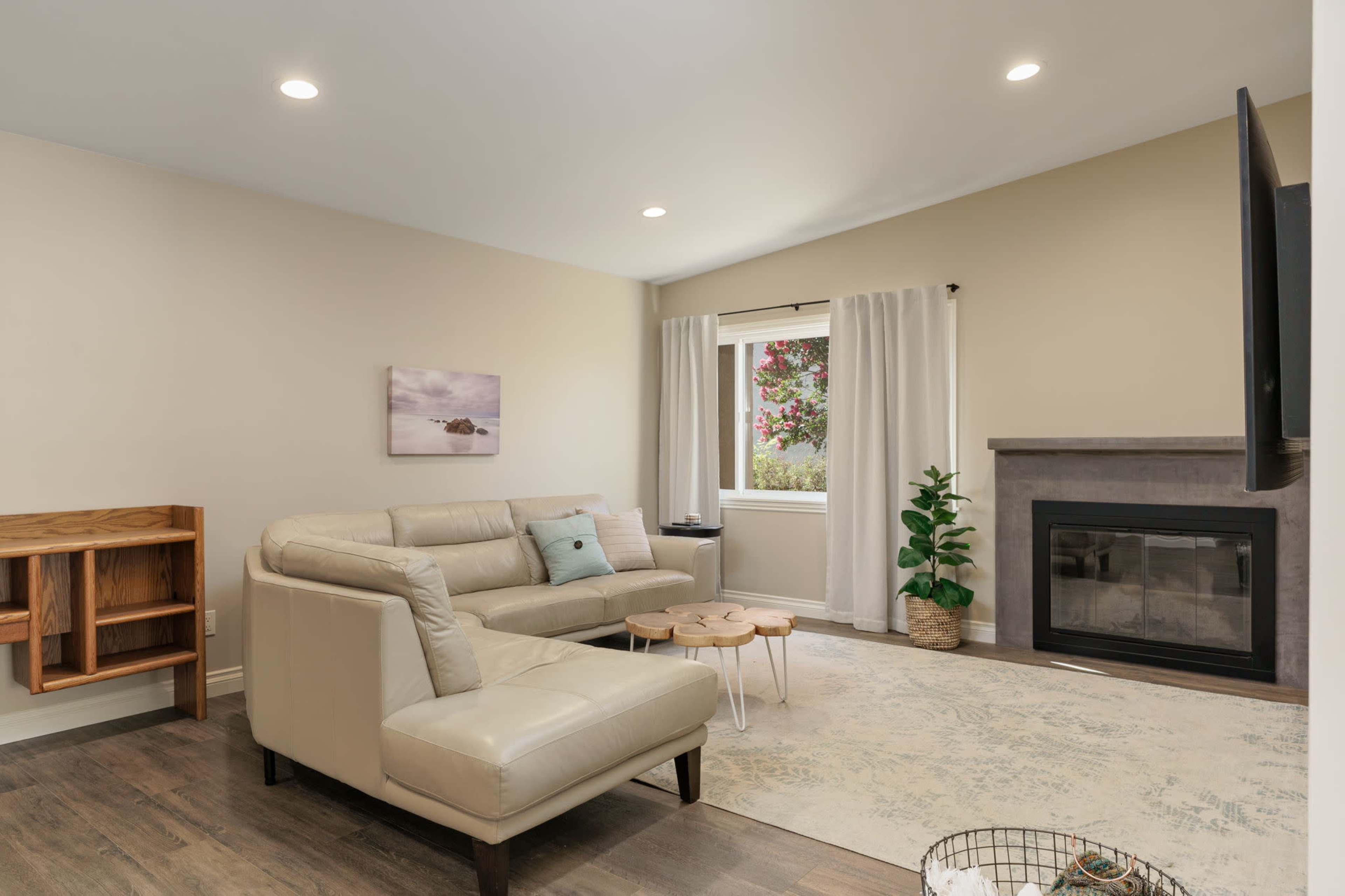 The image shows a modern living room with a light-colored sectional sofa, a small wooden side table, a fireplace, and a window with white curtains.