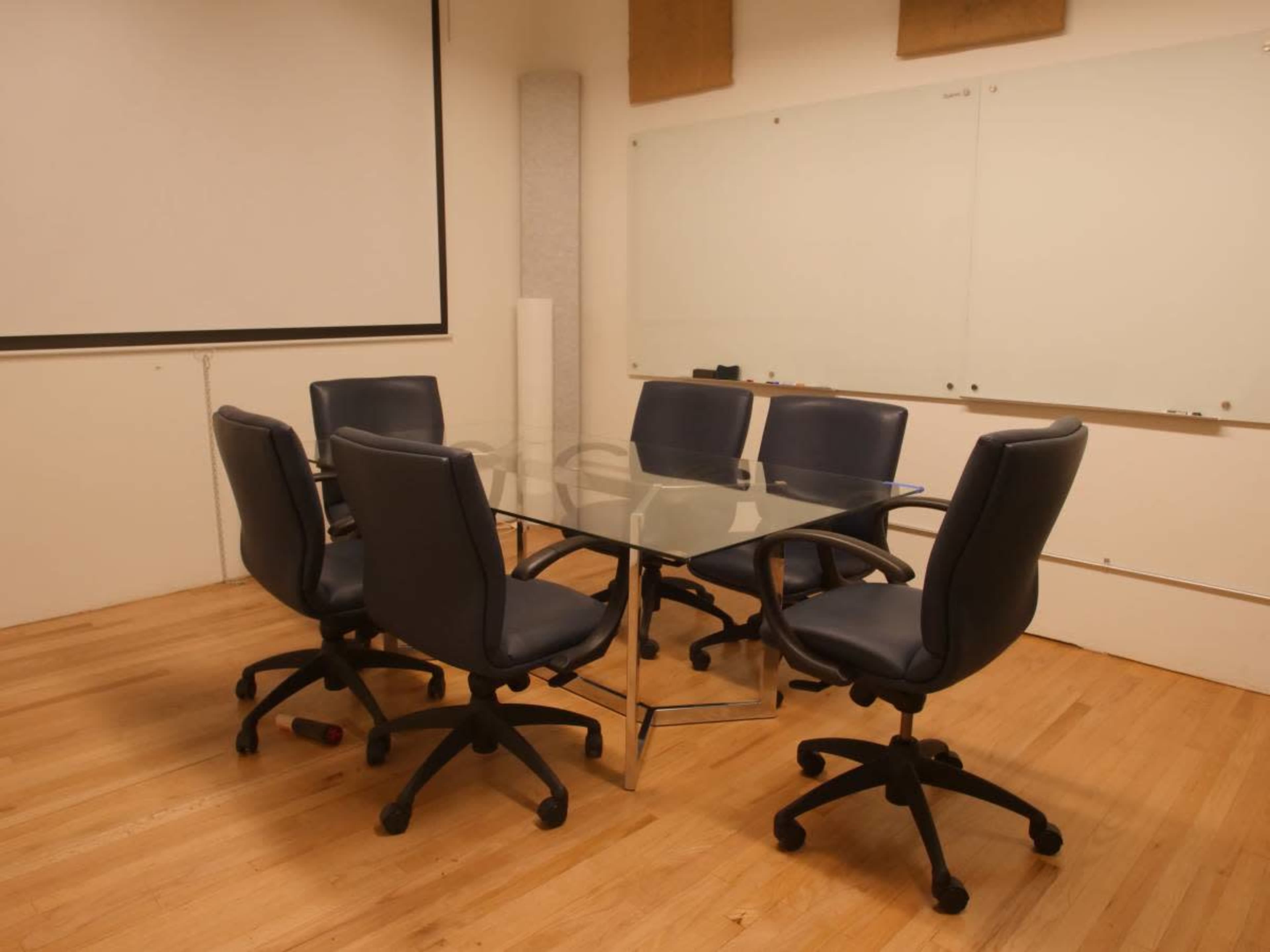 A conference room features a glass table surrounded by six blue office chairs on a wooden floor.