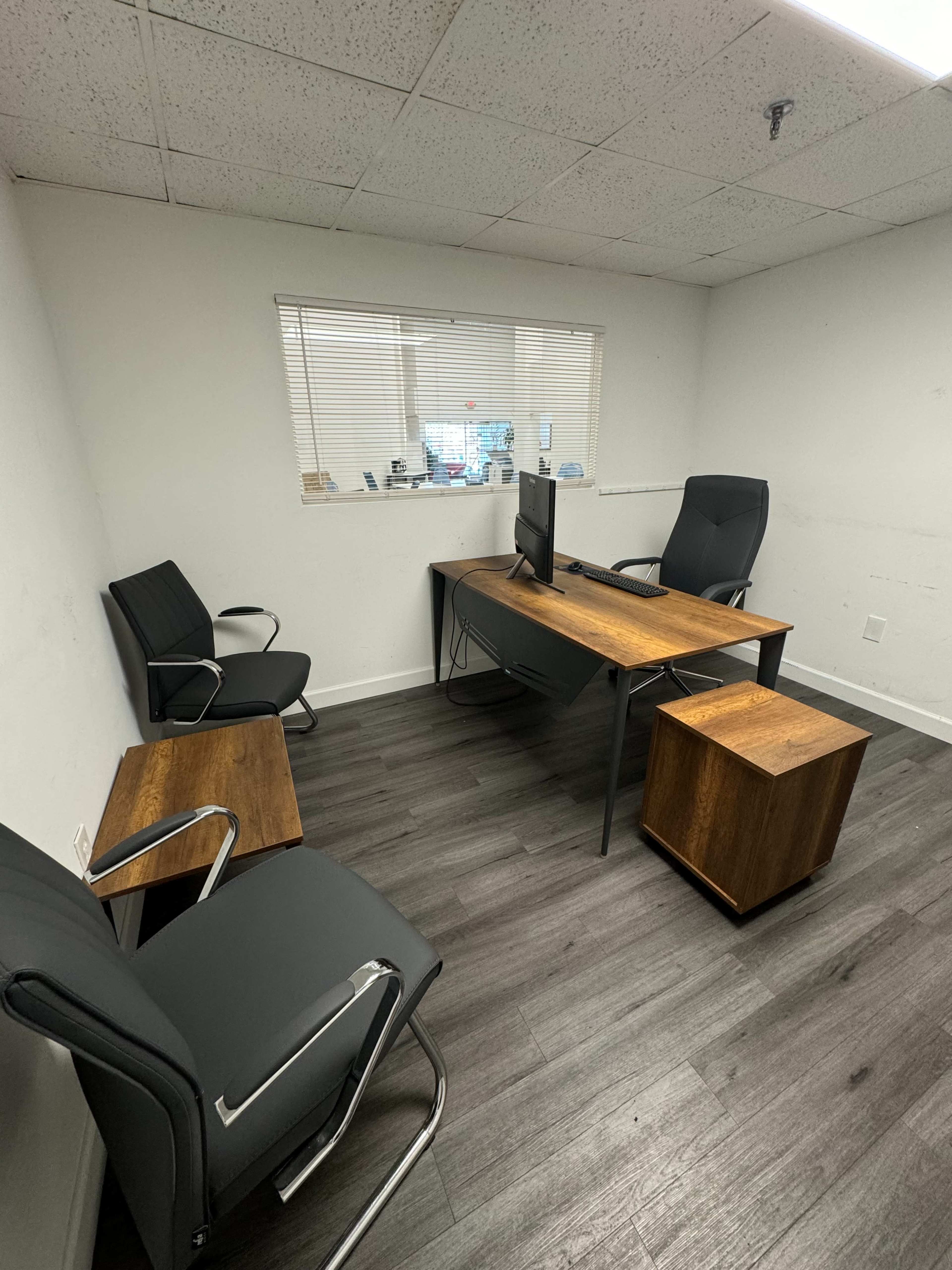 A minimalistic office space featuring a wooden desk with a monitor, two black chairs, and a small wooden accent table.