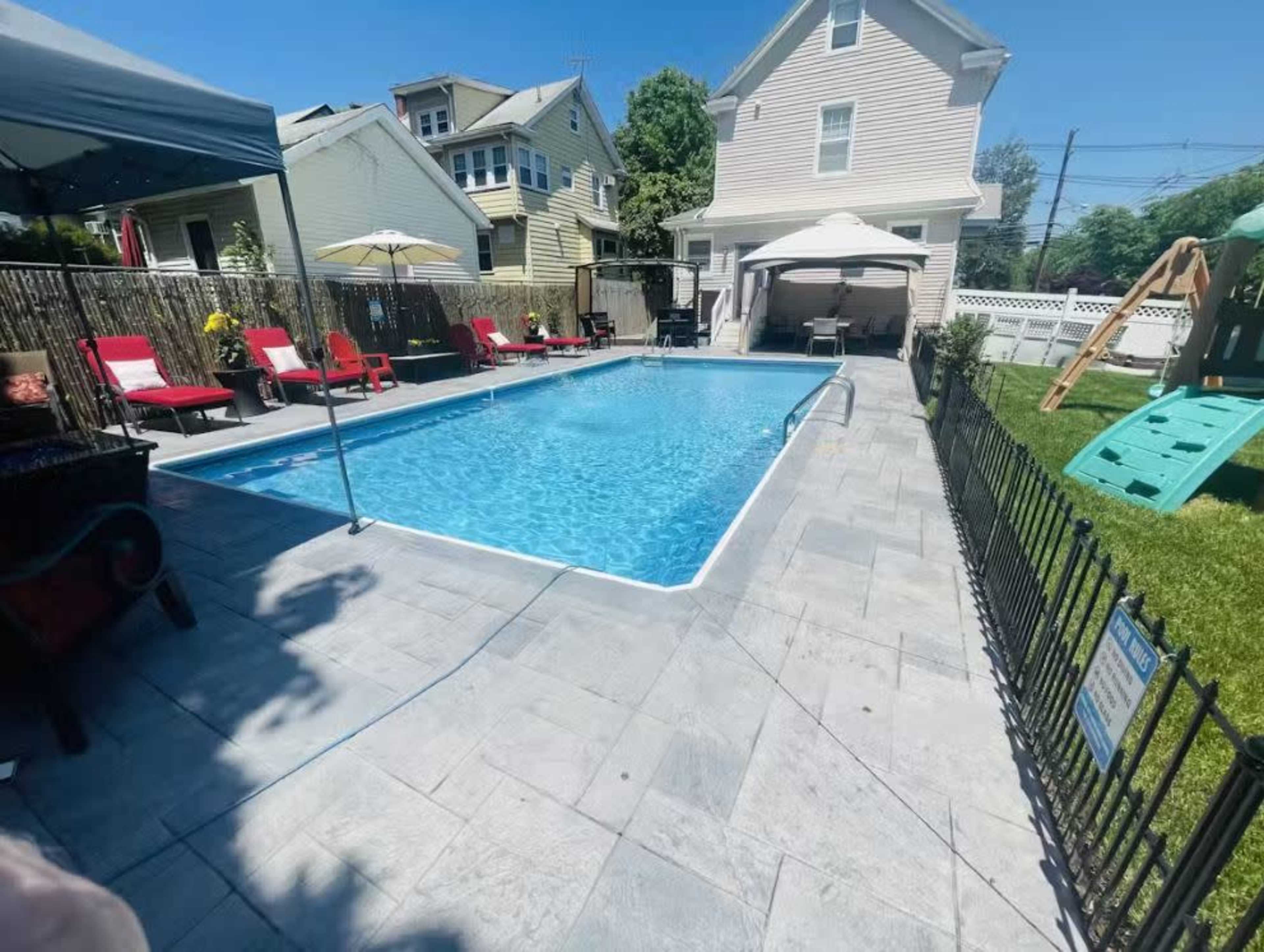 The image shows a rectangular swimming pool surrounded by patio furniture and a grassy area, with a house and a playset in the background.