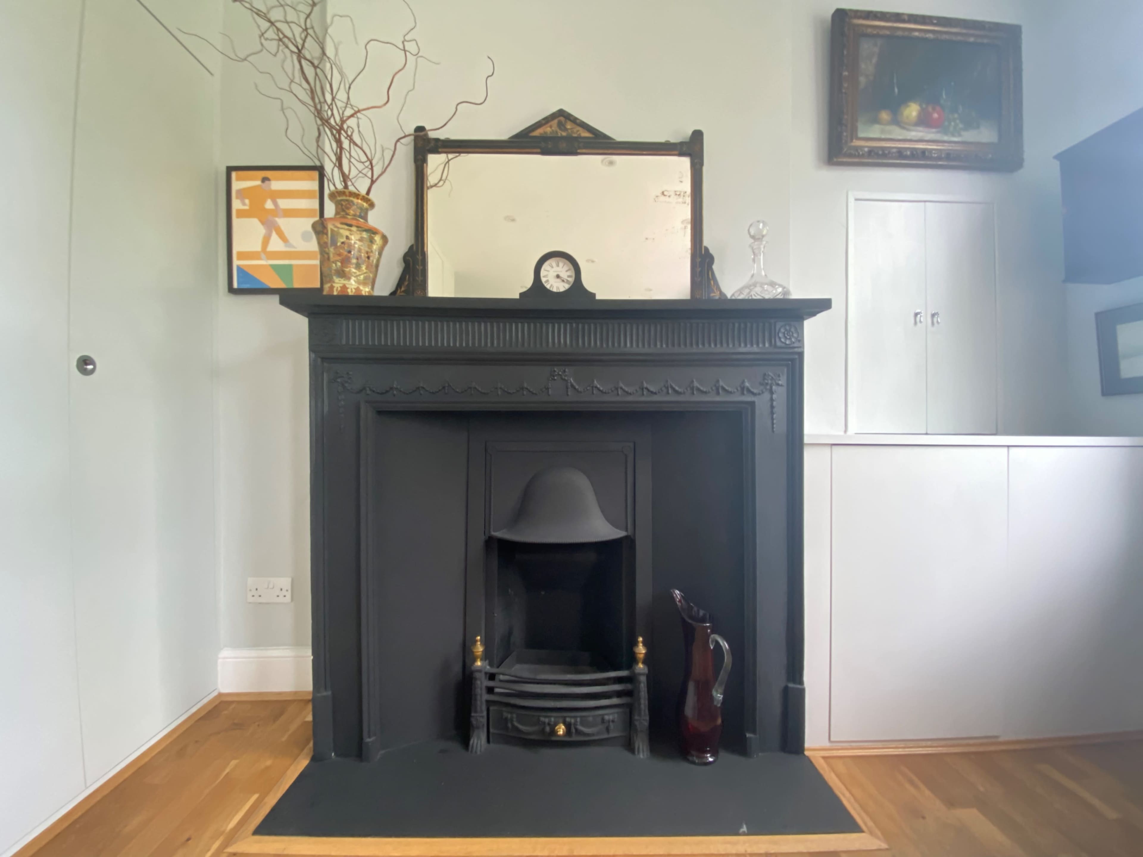 A black fireplace with a mirror and decorative items above it, set against a light-colored wall and a wooden floor.