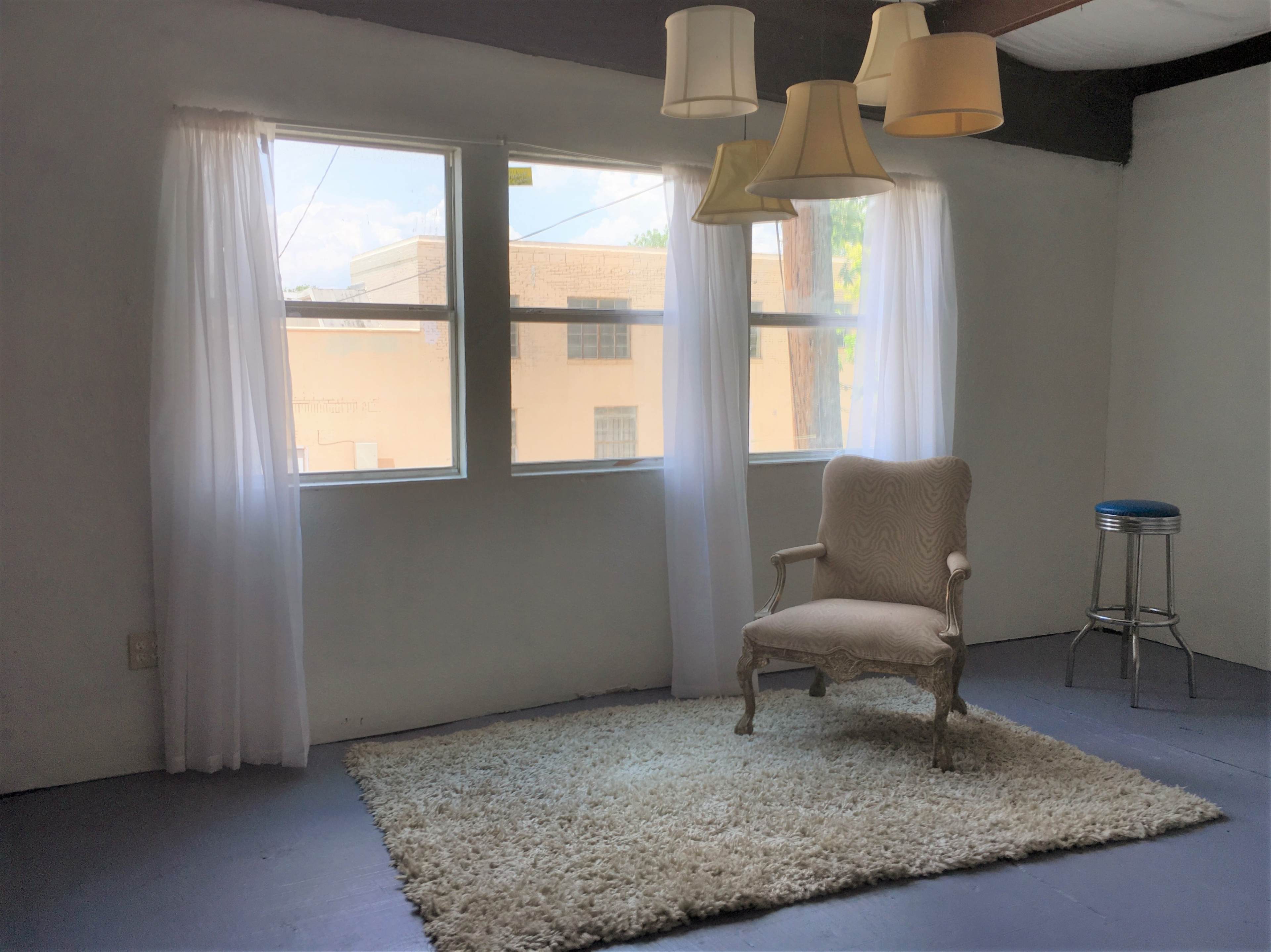 A decorated room features a plush rug, a vintage chair, and hanging light fixtures near large windows with sheer curtains.