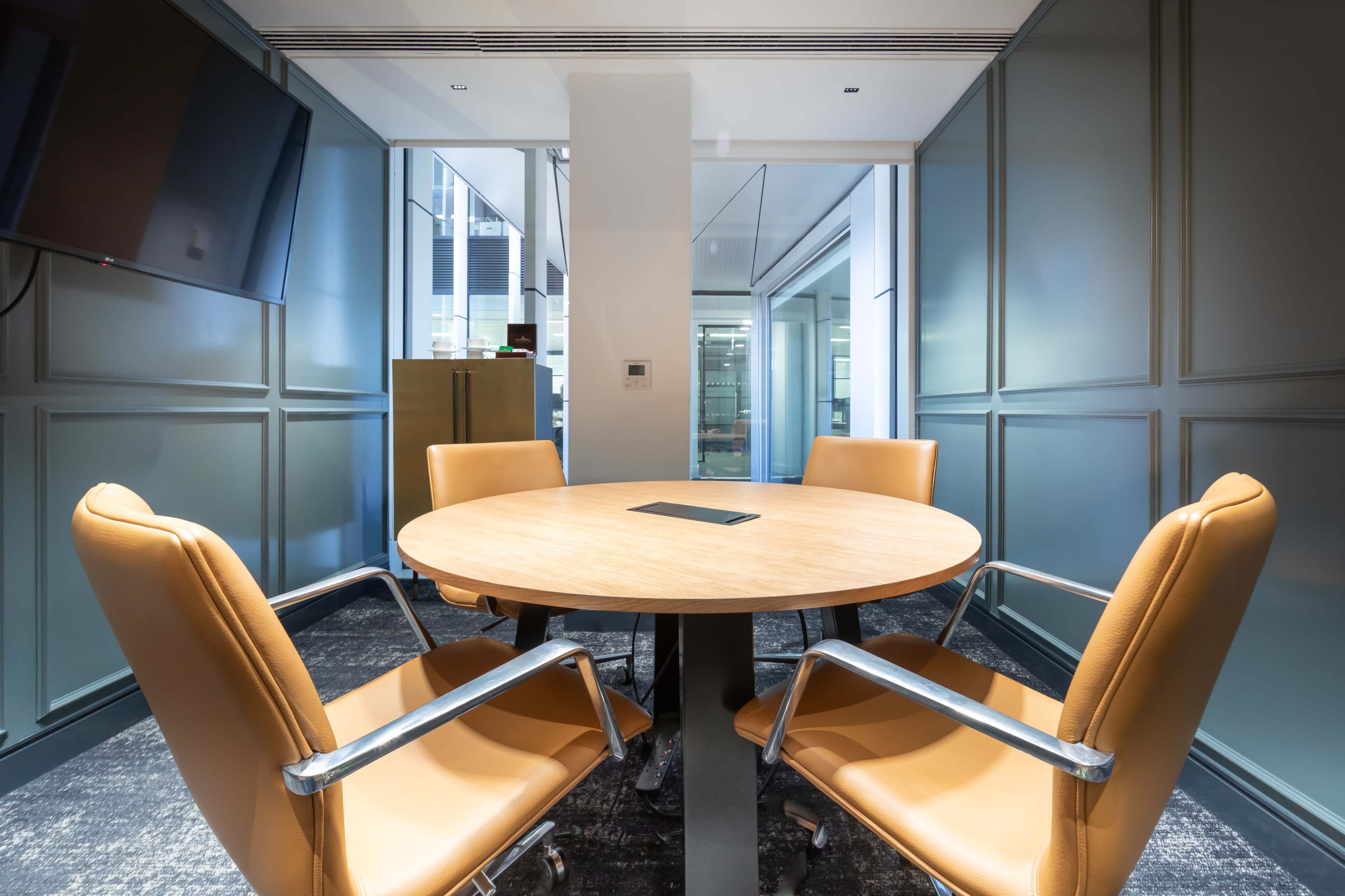 A modern conference room with a round wooden table and four leather chairs arranged around it, against a backdrop of glass walls.