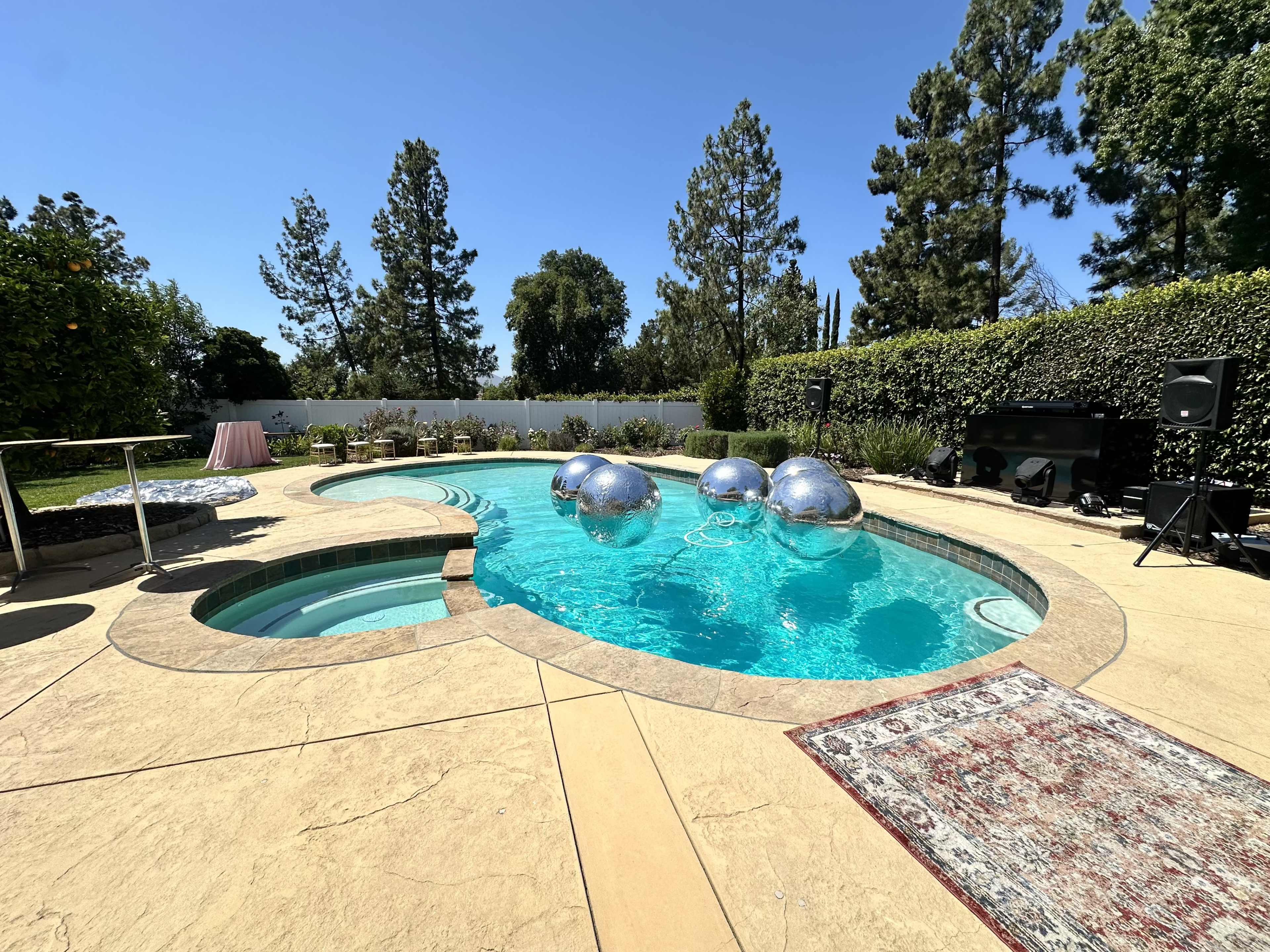 The image shows a backyard pool area with a large pool, a hot tub, and three reflective silver balls floating in the water, surrounded by trees and a DJ setup.