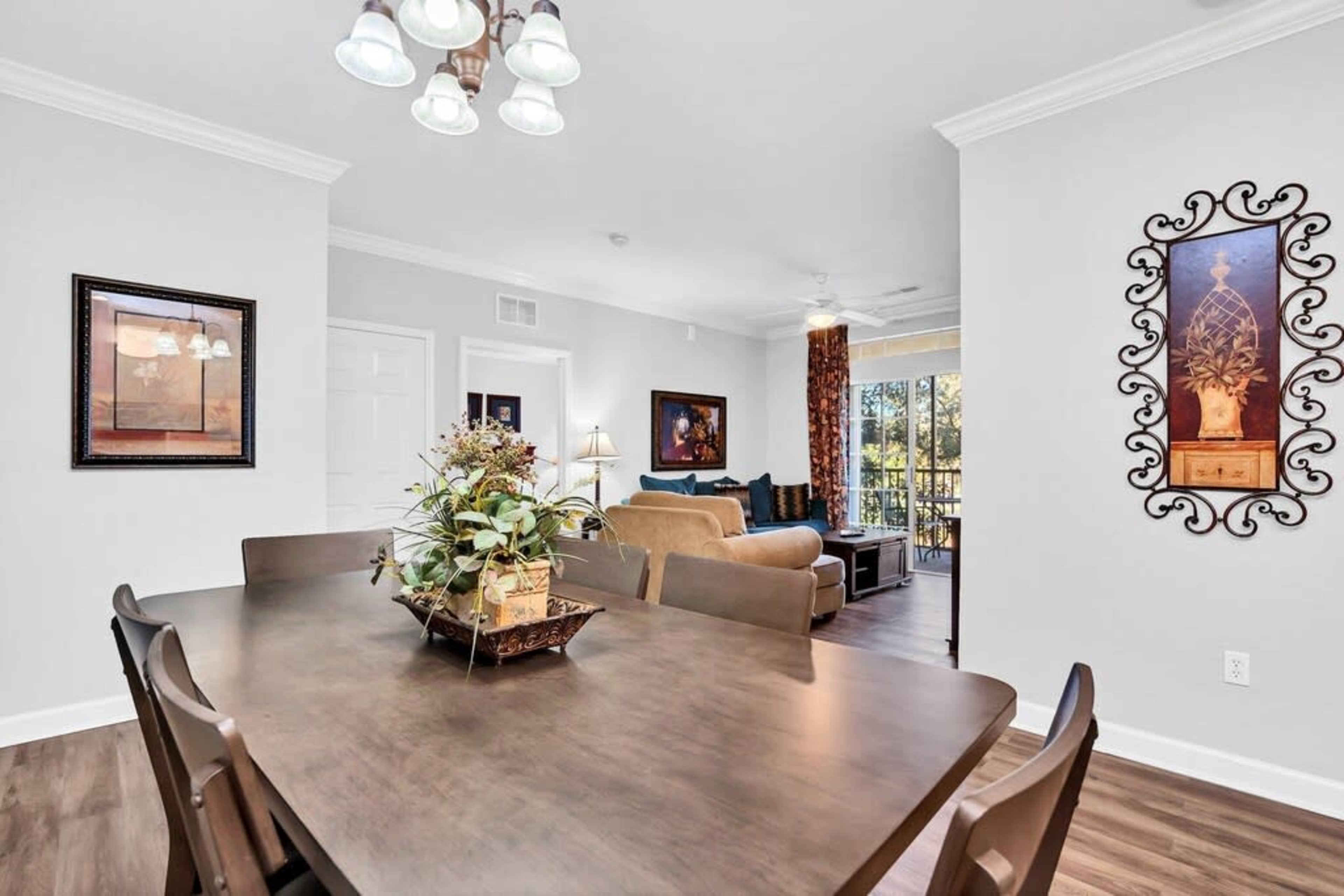 A dining area with a wooden table, decorative plants, and artwork, leading into a living room with a sofa and large windows.