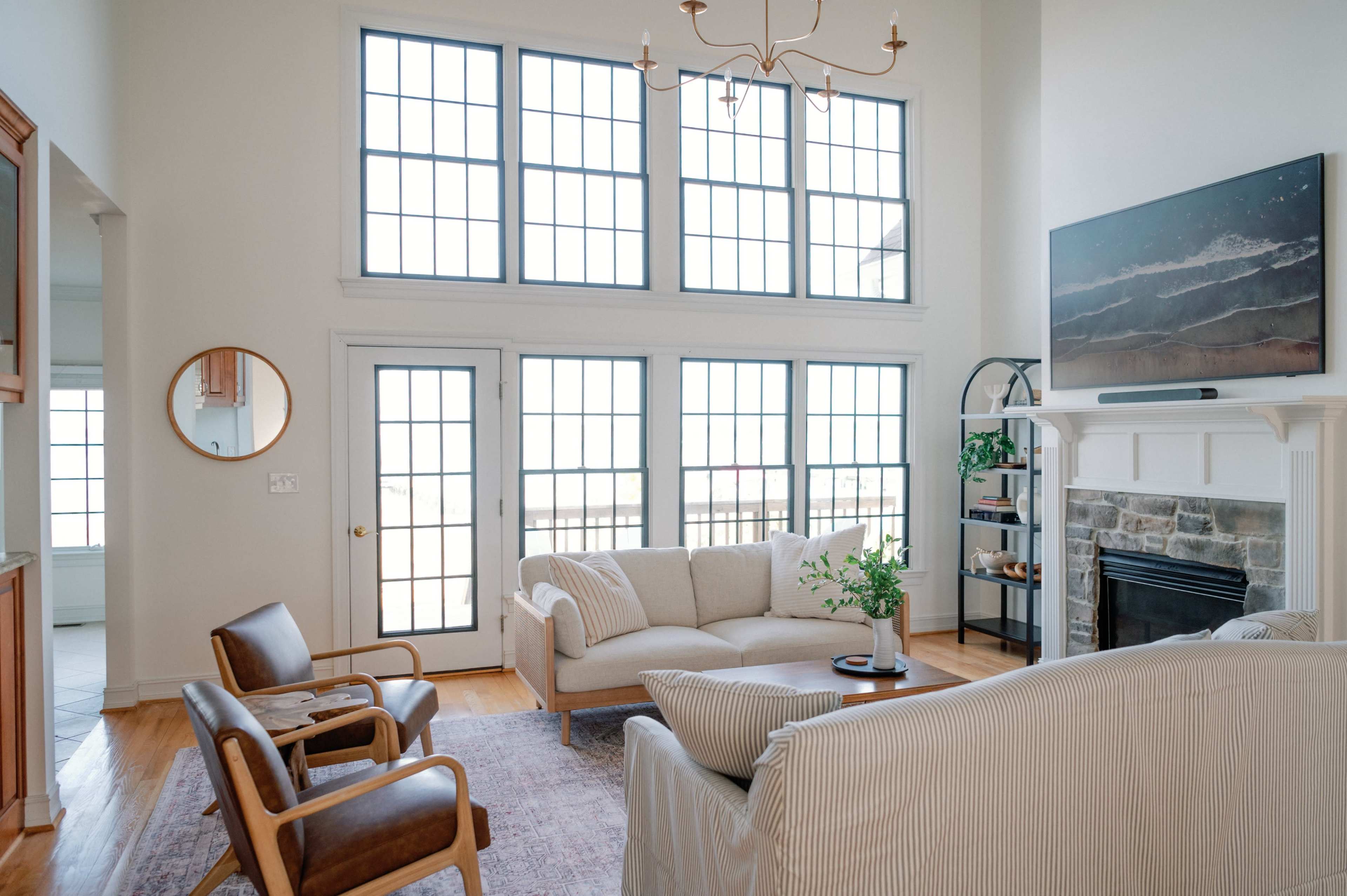 A spacious living room features large windows, a light-colored sofa, two armchairs, and a stone fireplace.